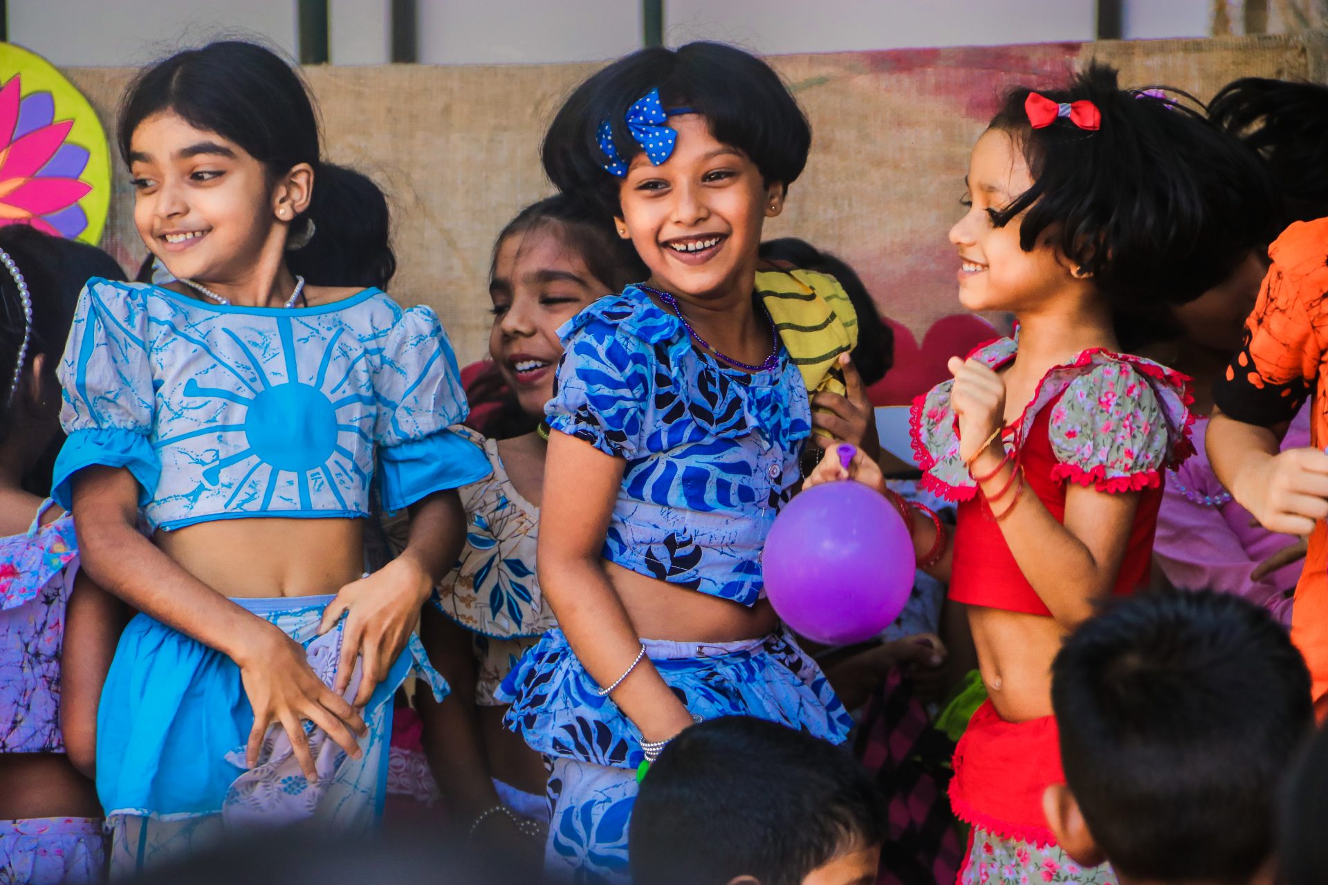 Children in colorful dresses smiling, holding purple balloon at indoor celebration.