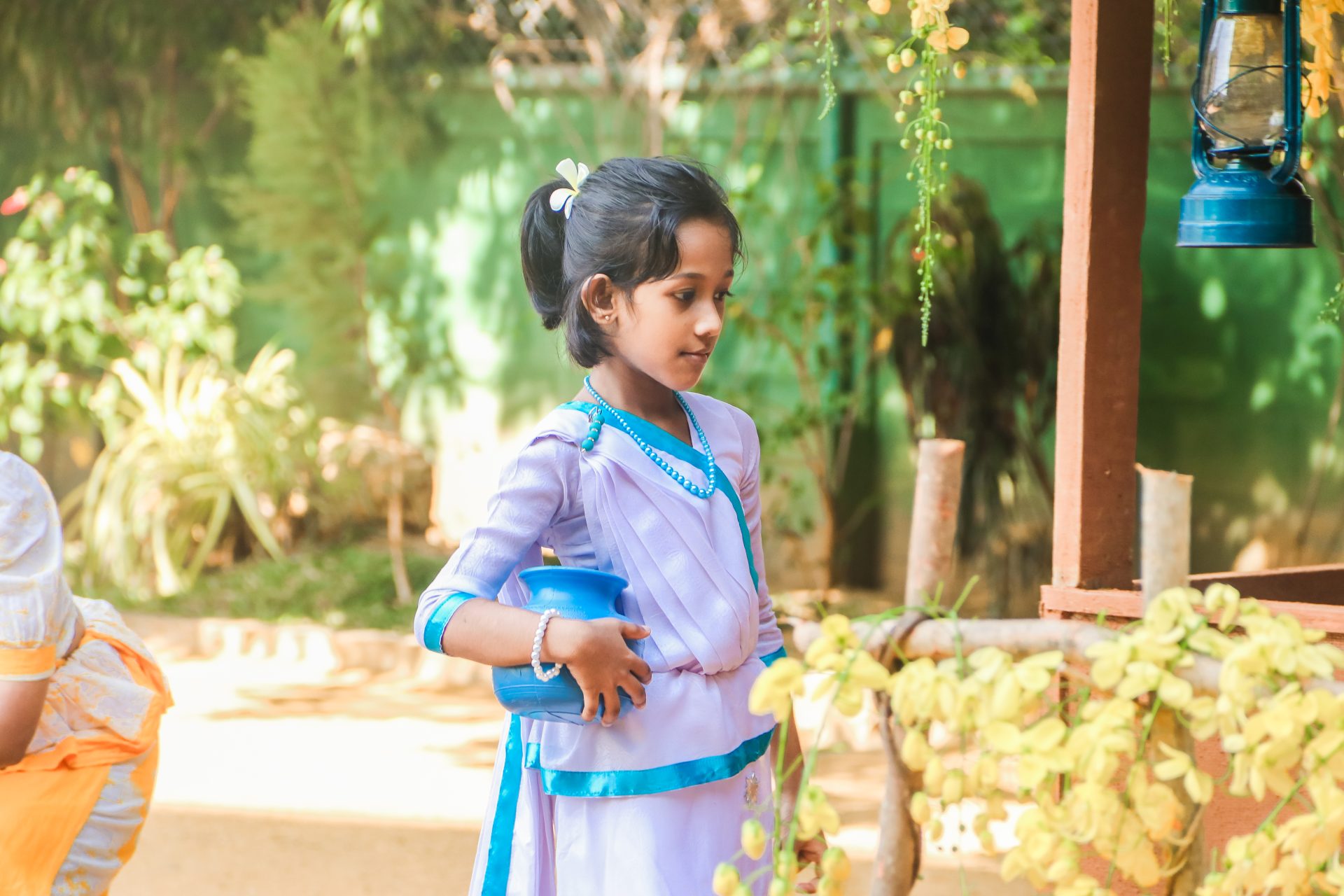 Young girl in traditional dress holding a blue pot in a vibrant garden.