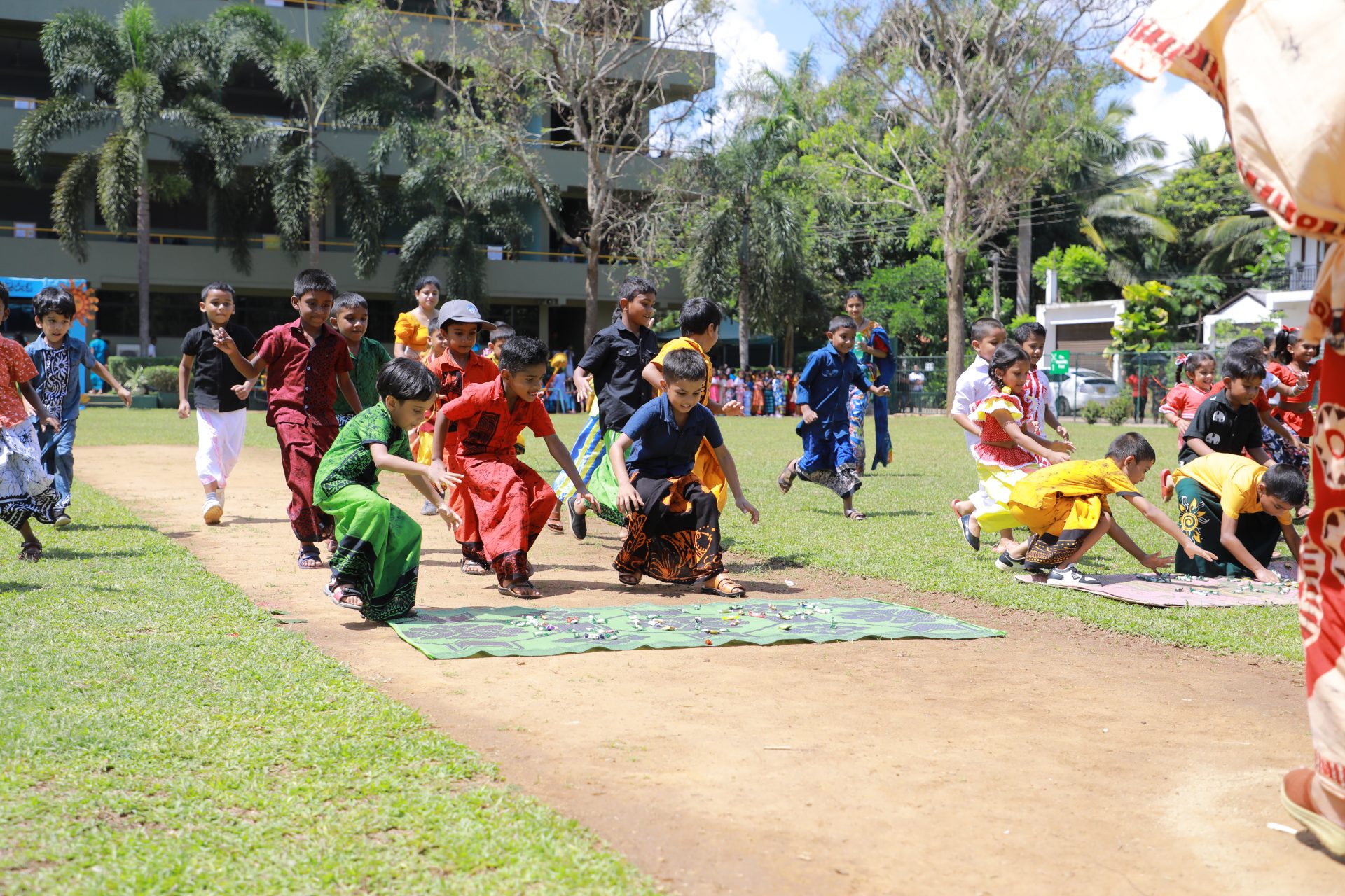 Children in traditional attire racing on a sunny grassy field, surrounded by cheering peers.