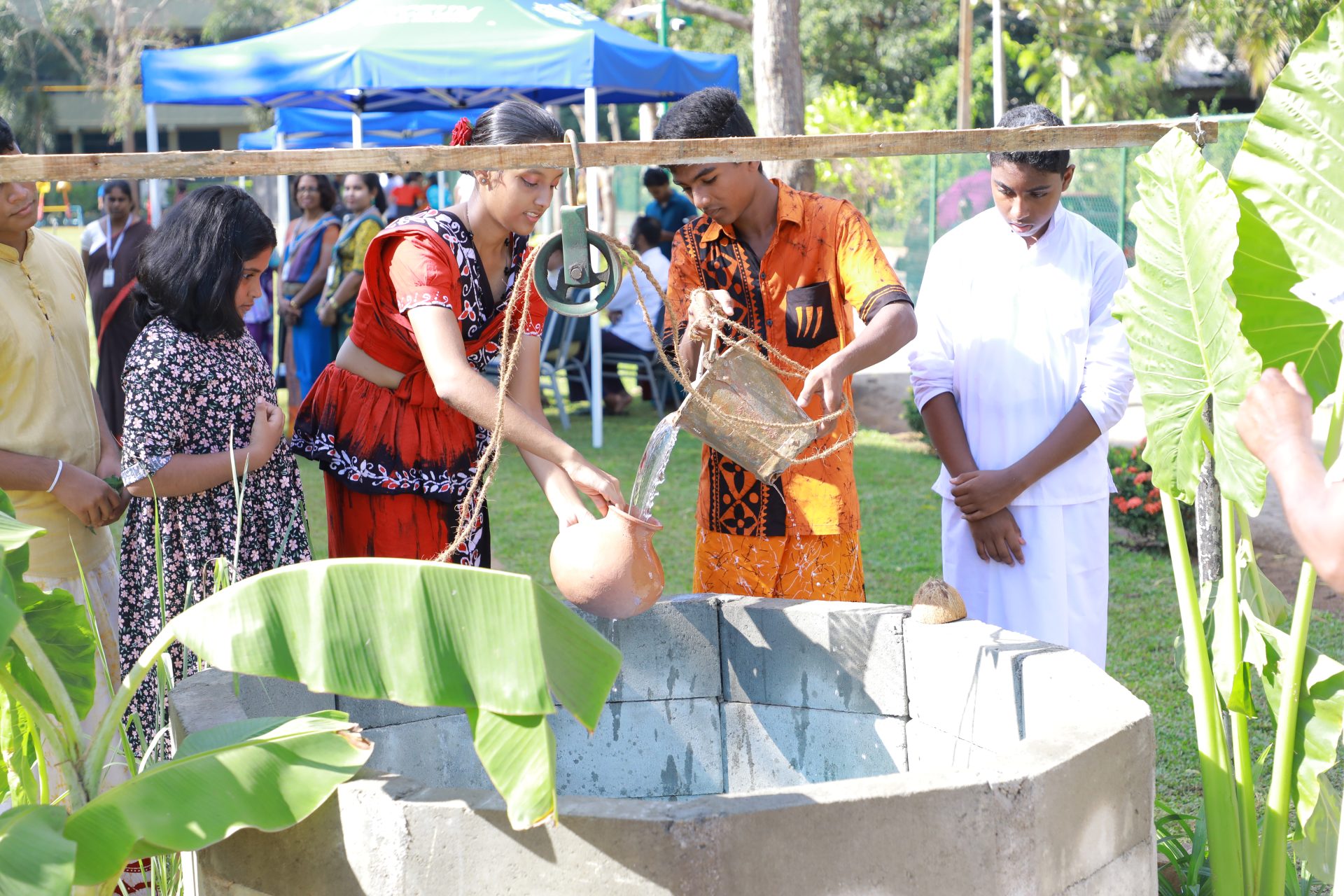 Community members engage in traditional water ceremony at a well, surrounded by greenery.