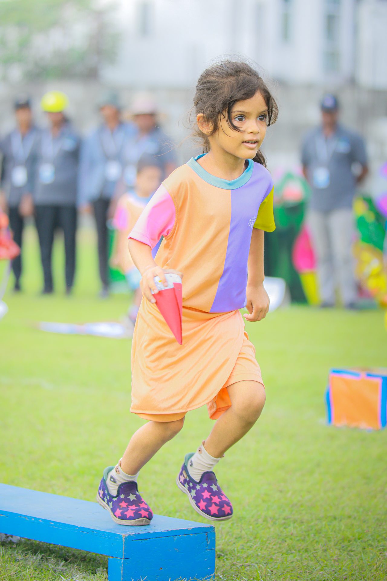 Young girl in colorful outfit on obstacle course, holding red cone.