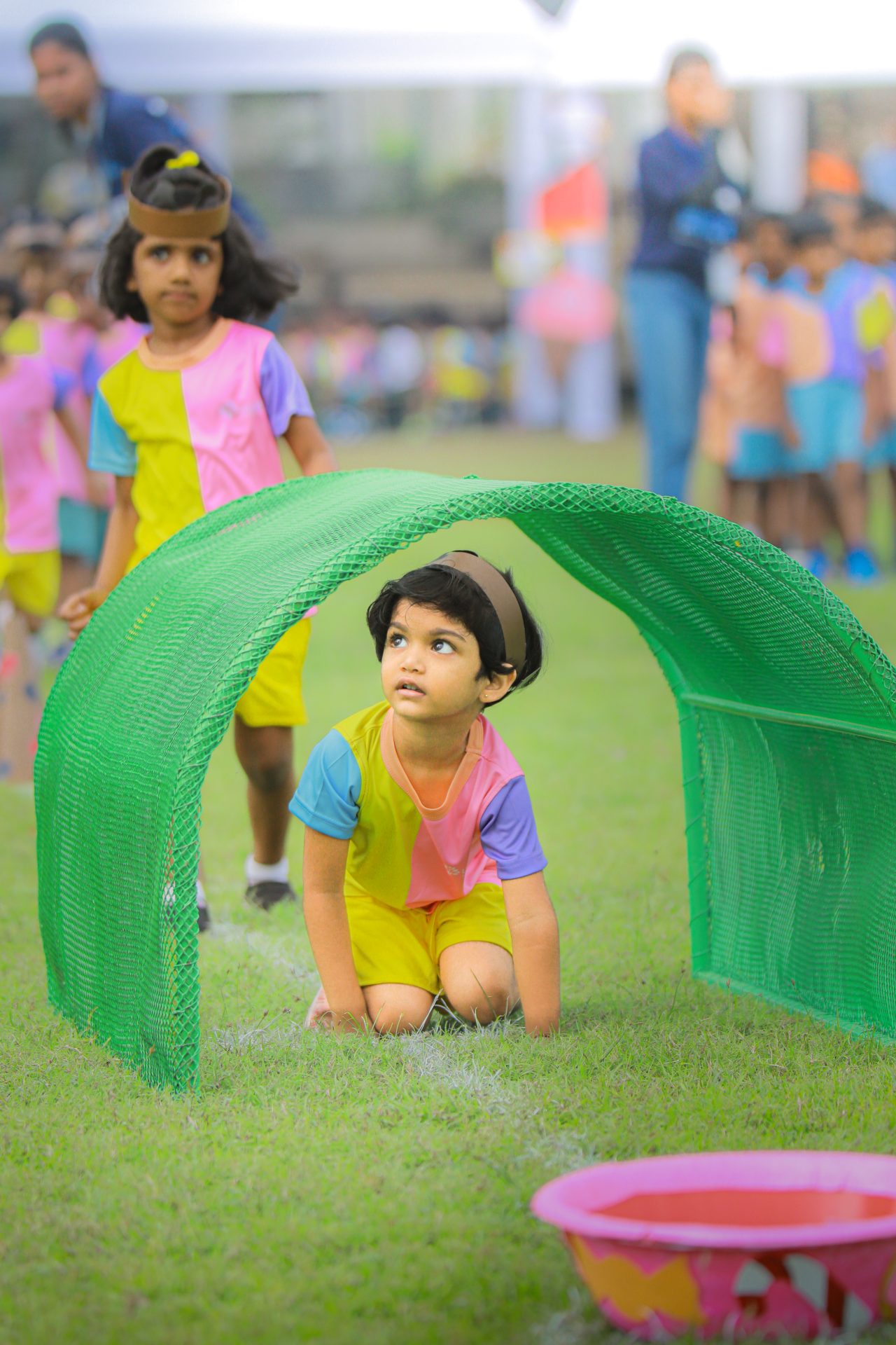 Children crawling through a green tunnel at a lively school event on a grassy field.