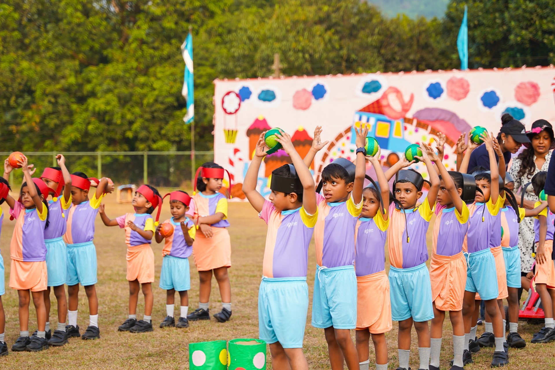 Children playing outdoor games in colorful uniforms with a festive backdrop and greenery.