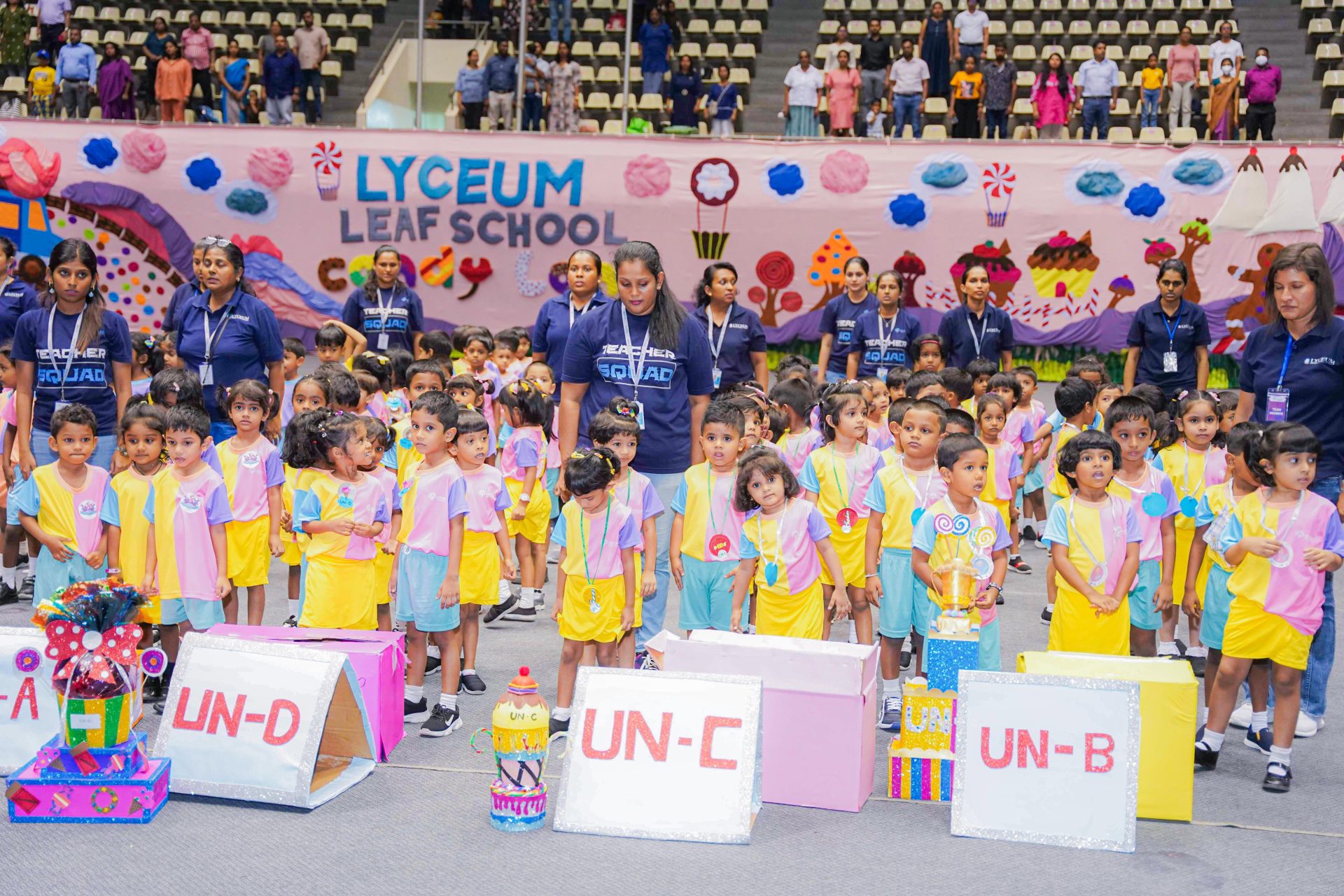 Children in colorful uniforms at Lyceum Leaf School event with teachers and festive backdrop.