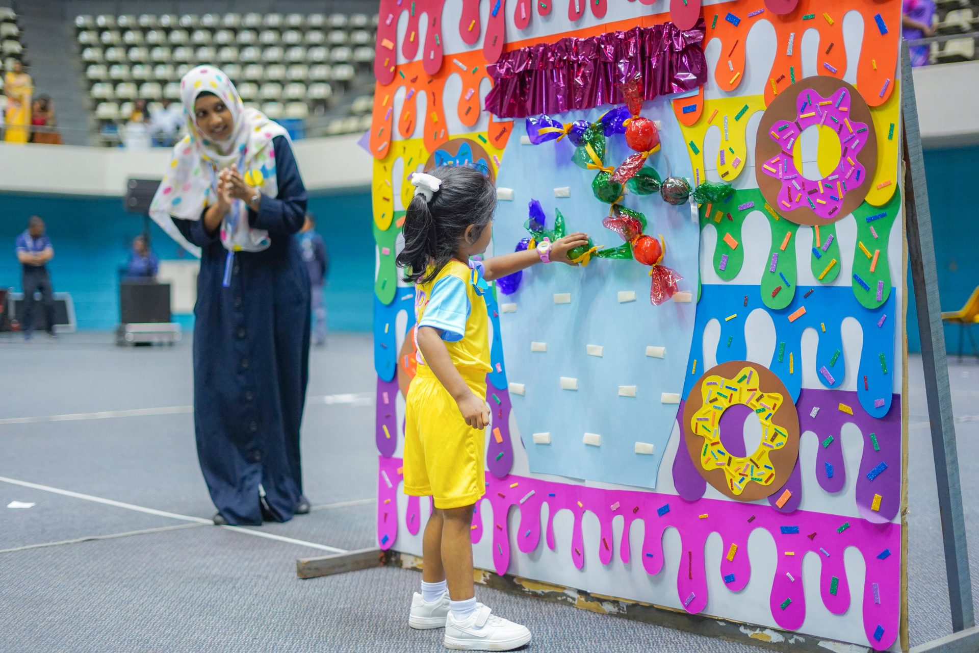 Young girl interacts with colorful display at indoor fair, woman observes.