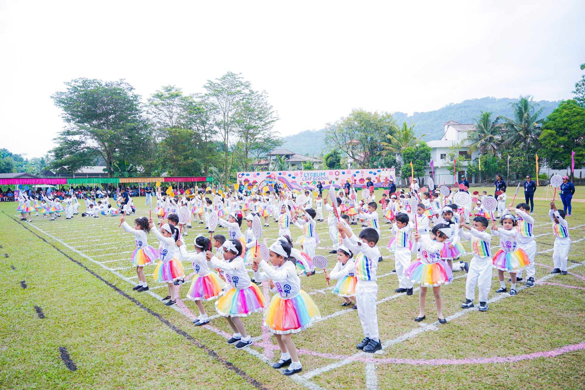 Children perform a colorful dance on a green field under a partly cloudy sky.