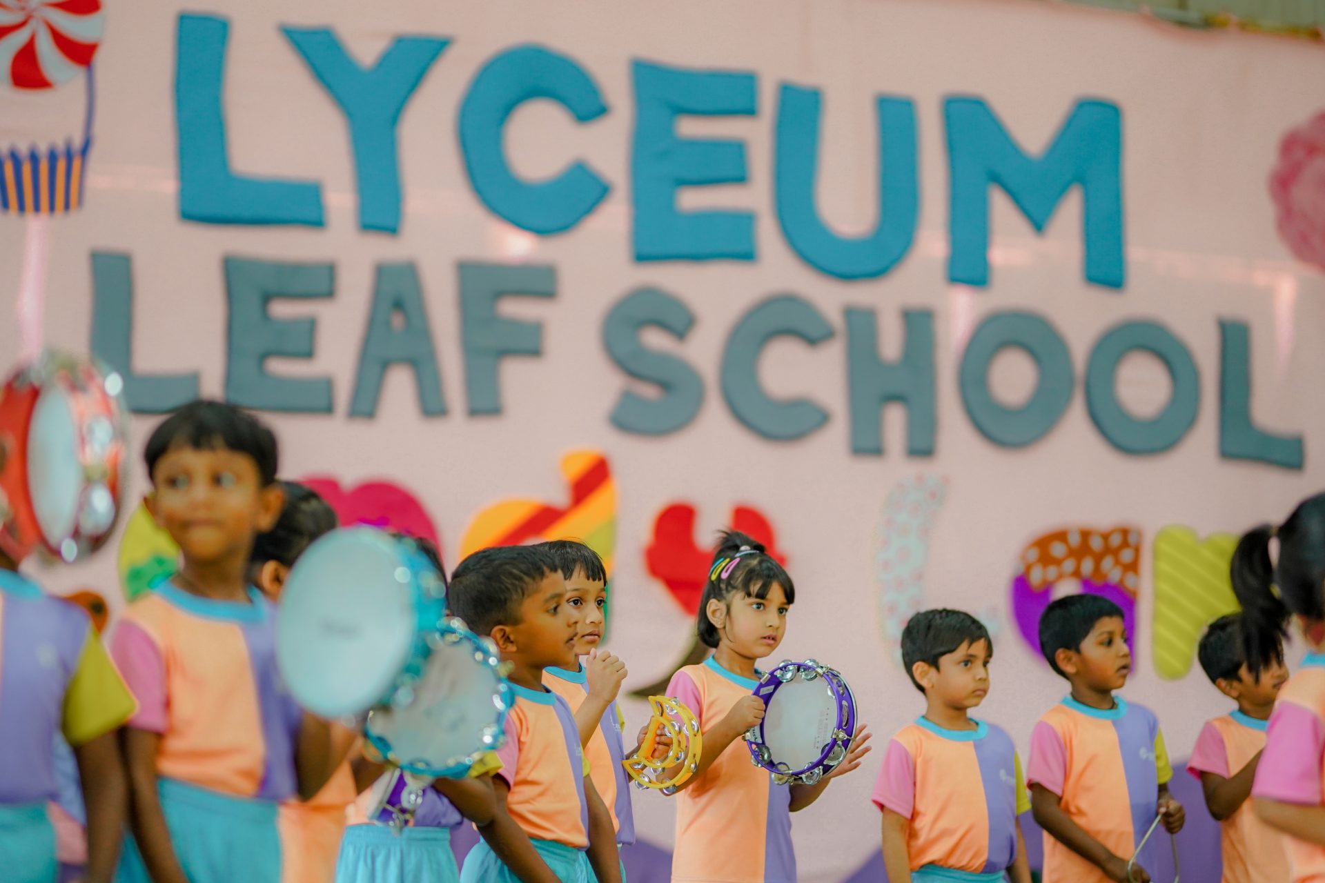 Children performing tambourine dance at Lyceum Leaf School event, vibrant backdrop in background.