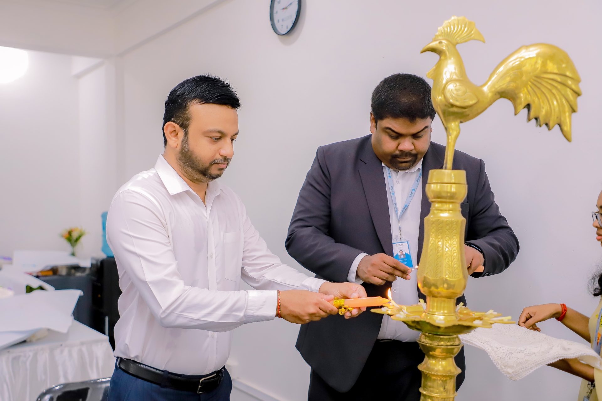 Men in formal attire light a traditional brass oil lamp in a ceremonial event.