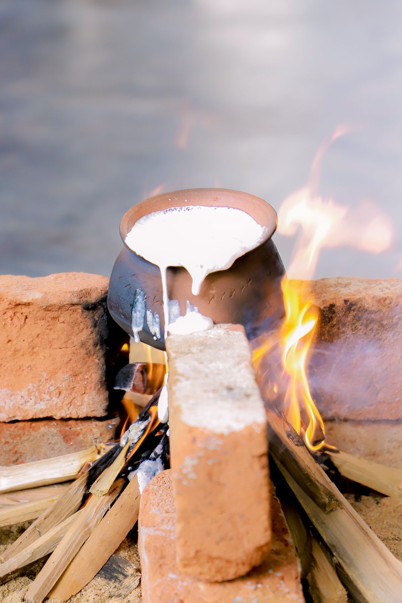 Boiling milk overflows from a metal pot on a brick stove with vibrant flames.