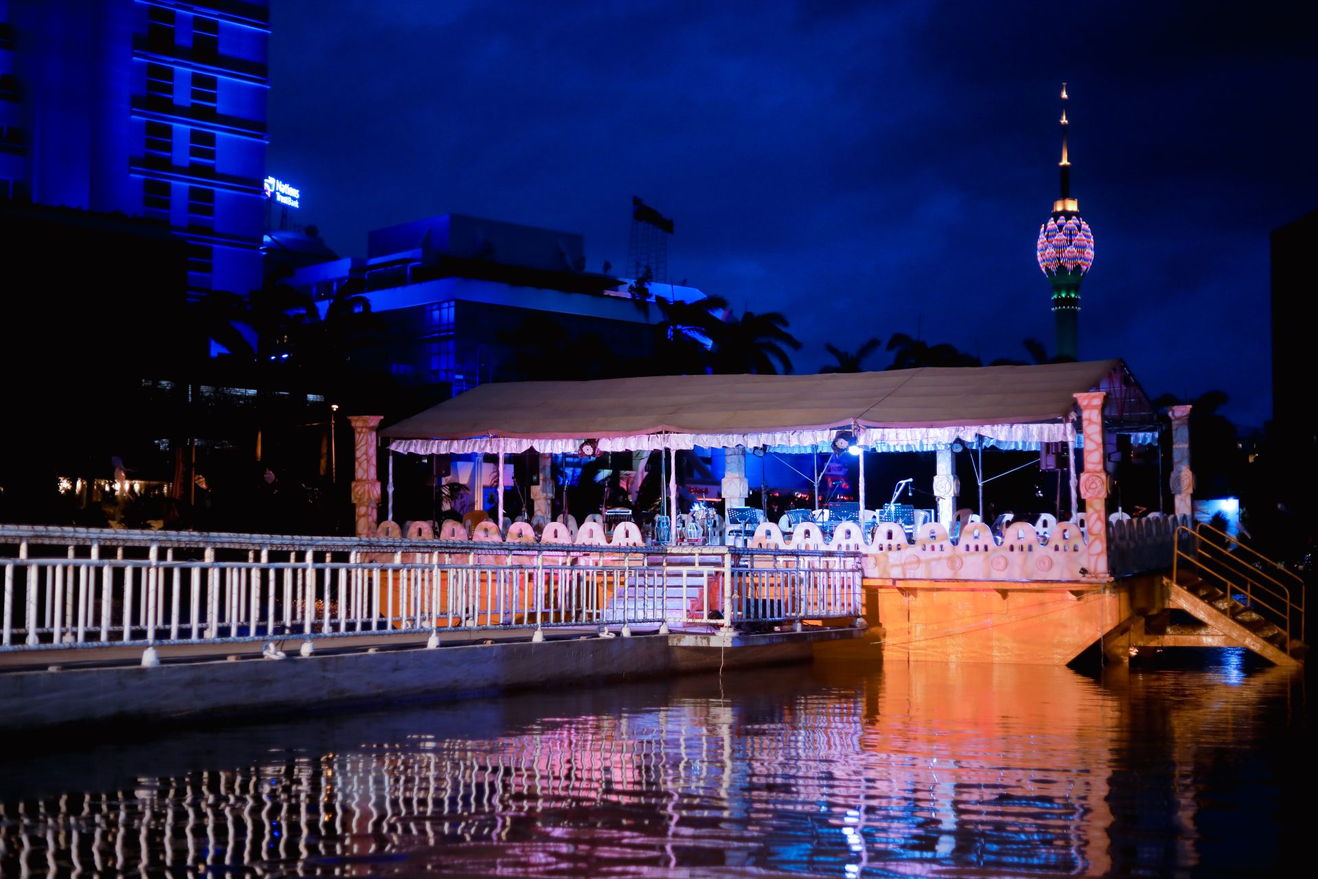 Serene waterfront evening with illuminated pavilion, cityscape, and lotus-shaped tower reflections.