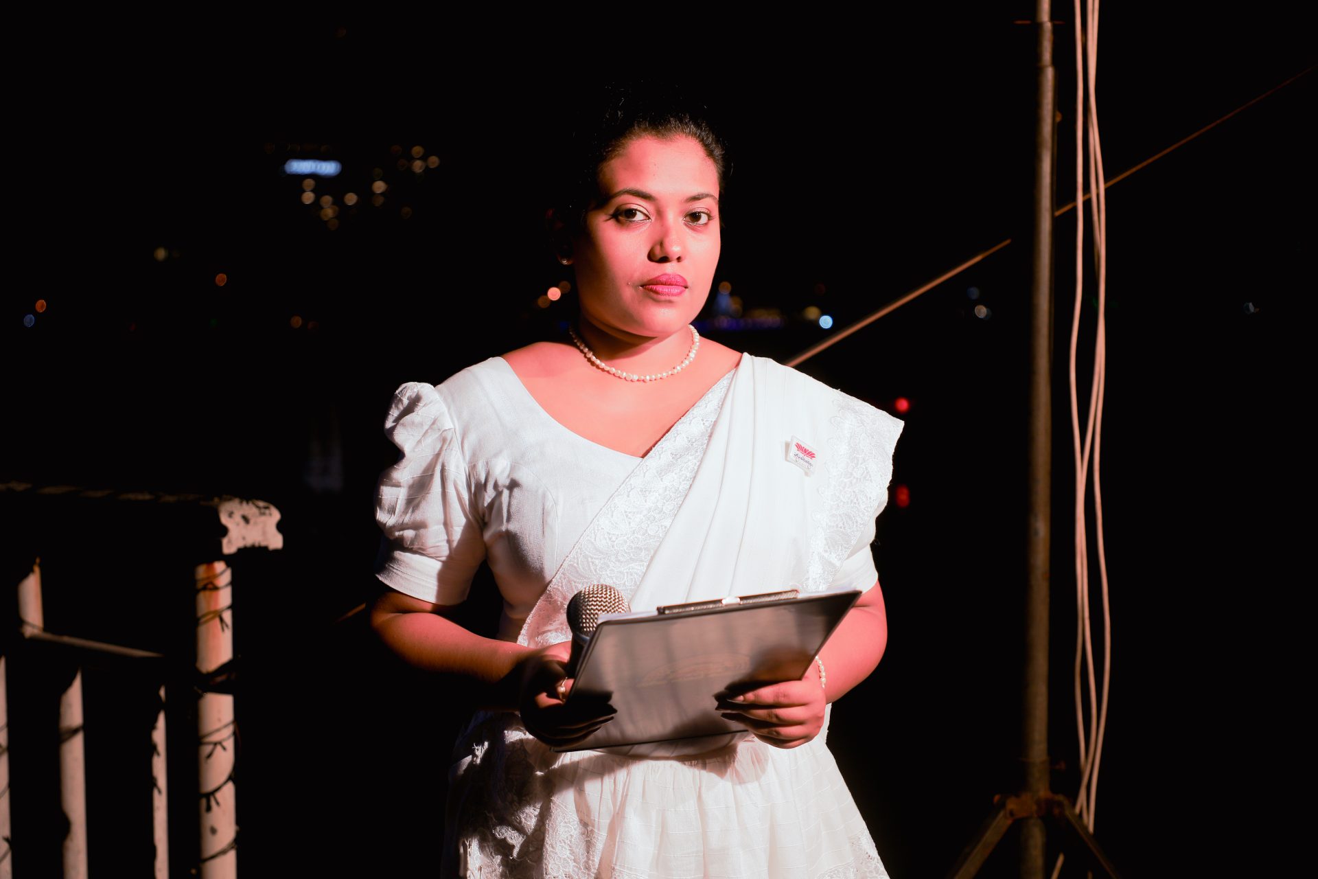 Woman in white saree holding microphone and clipboard at nighttime event.