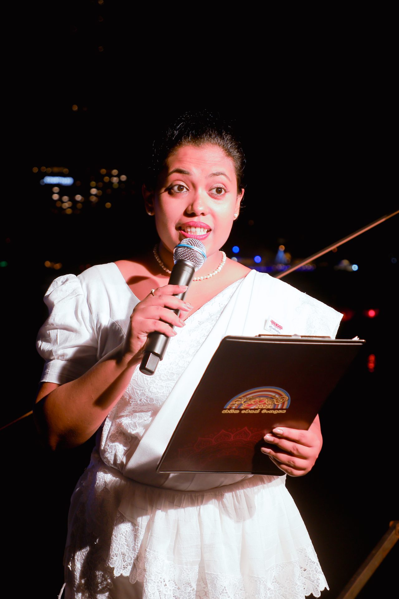Woman speaking confidently with microphone, wearing traditional attire, in spotlight against dark background.