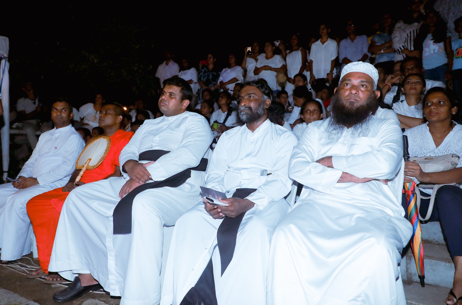 Interfaith leaders at a nighttime ceremony, diverse crowd in the background.