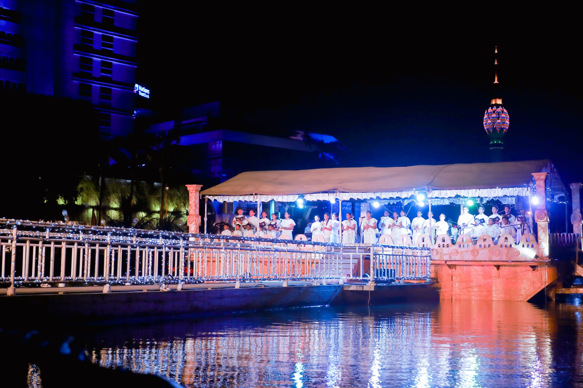 Illuminated floating platform night ceremony with festive lights and urban skyline.