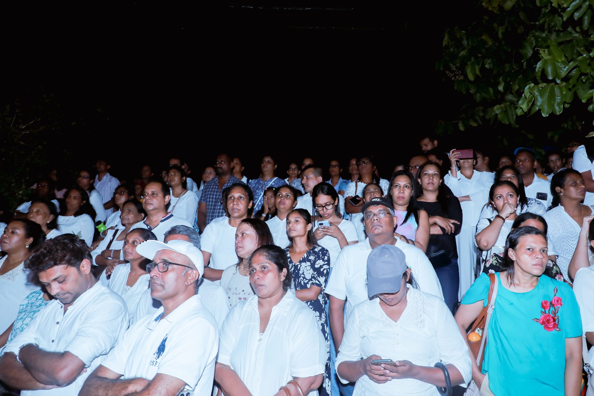 Diverse nighttime crowd in white attire, focused and solemn, some using smartphones.