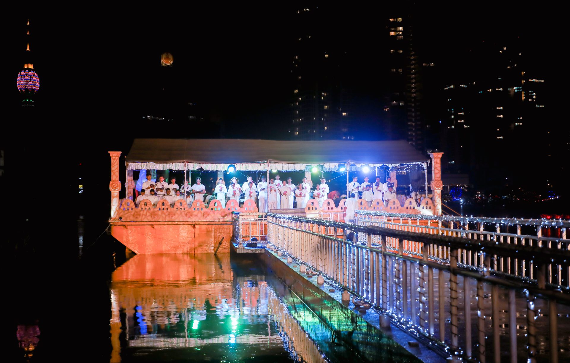 Nighttime floating ceremony on decorated boat, cityscape backdrop, festive lights reflect on water.