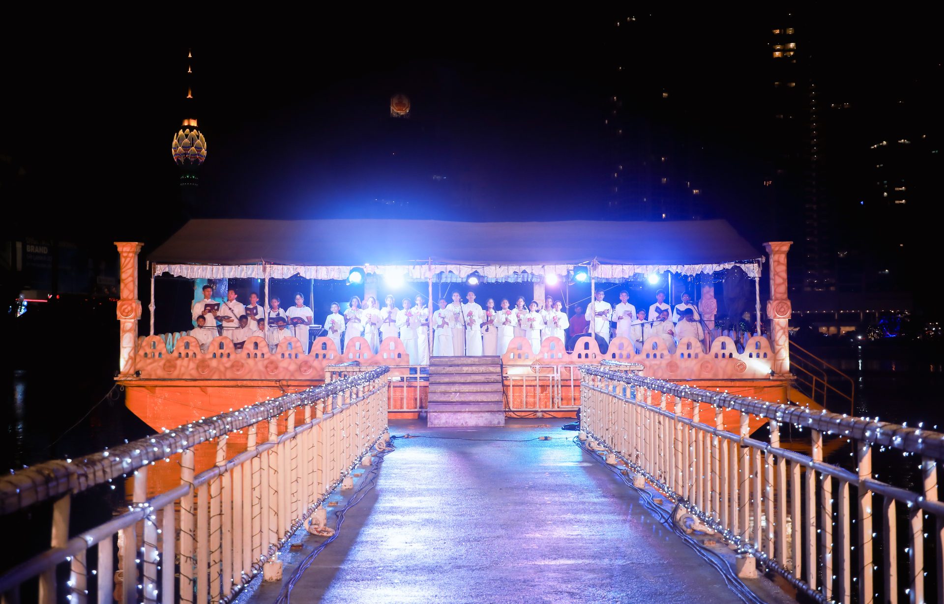 Festive night performance on ornately decorated floating platform with city skyline backdrop.