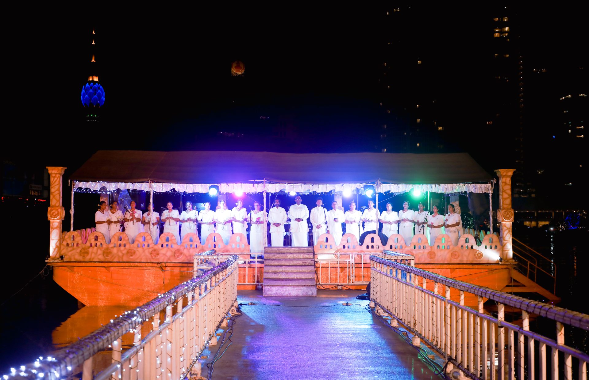 Floating night ceremony with individuals in white robes, ornate platform, and festive lights.