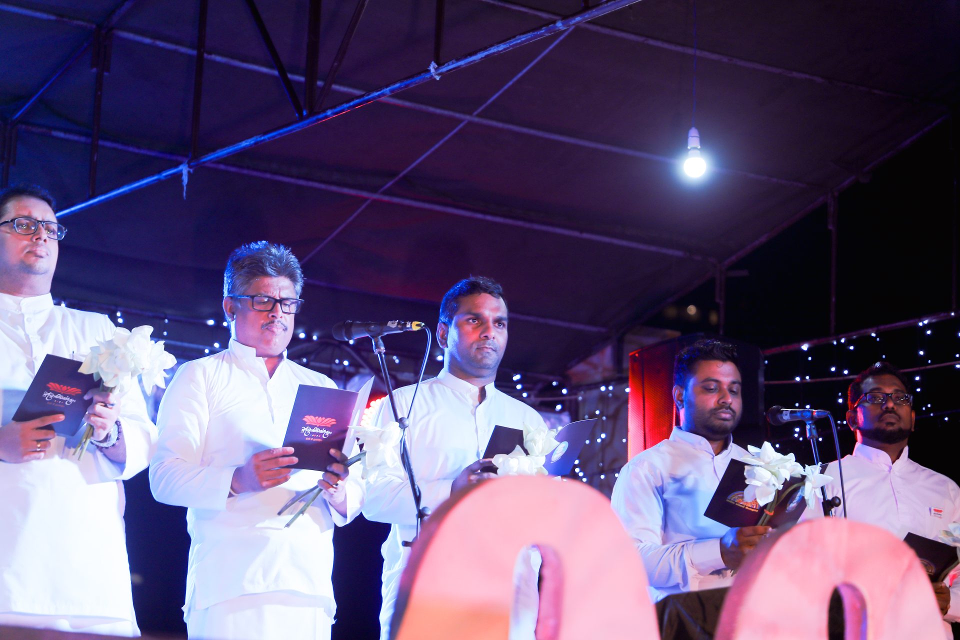 Formal ceremony with men in white attire holding booklets and flowers on stage.