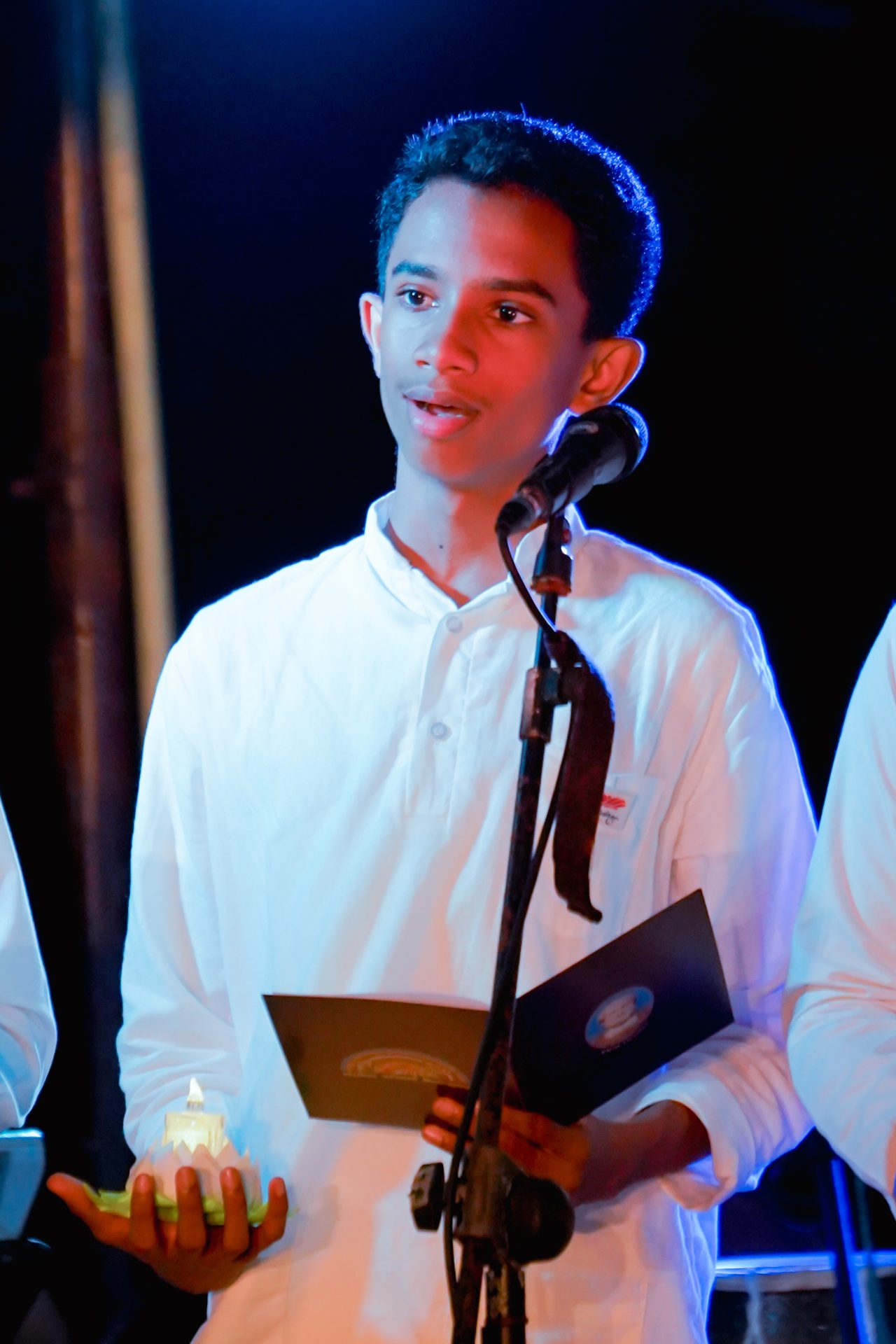 Young boy speaking at formal event, holding folder, under blue and red lighting.