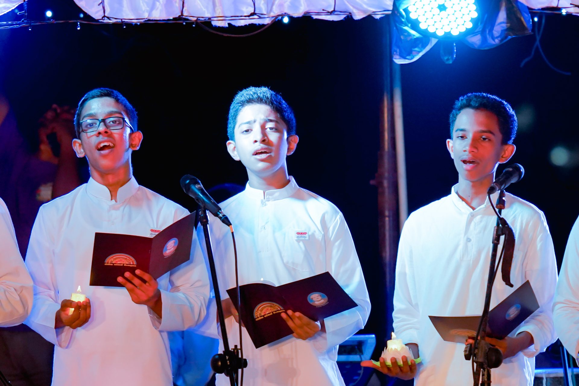 Young men in white traditional attire singing in choir with candles and booklets.