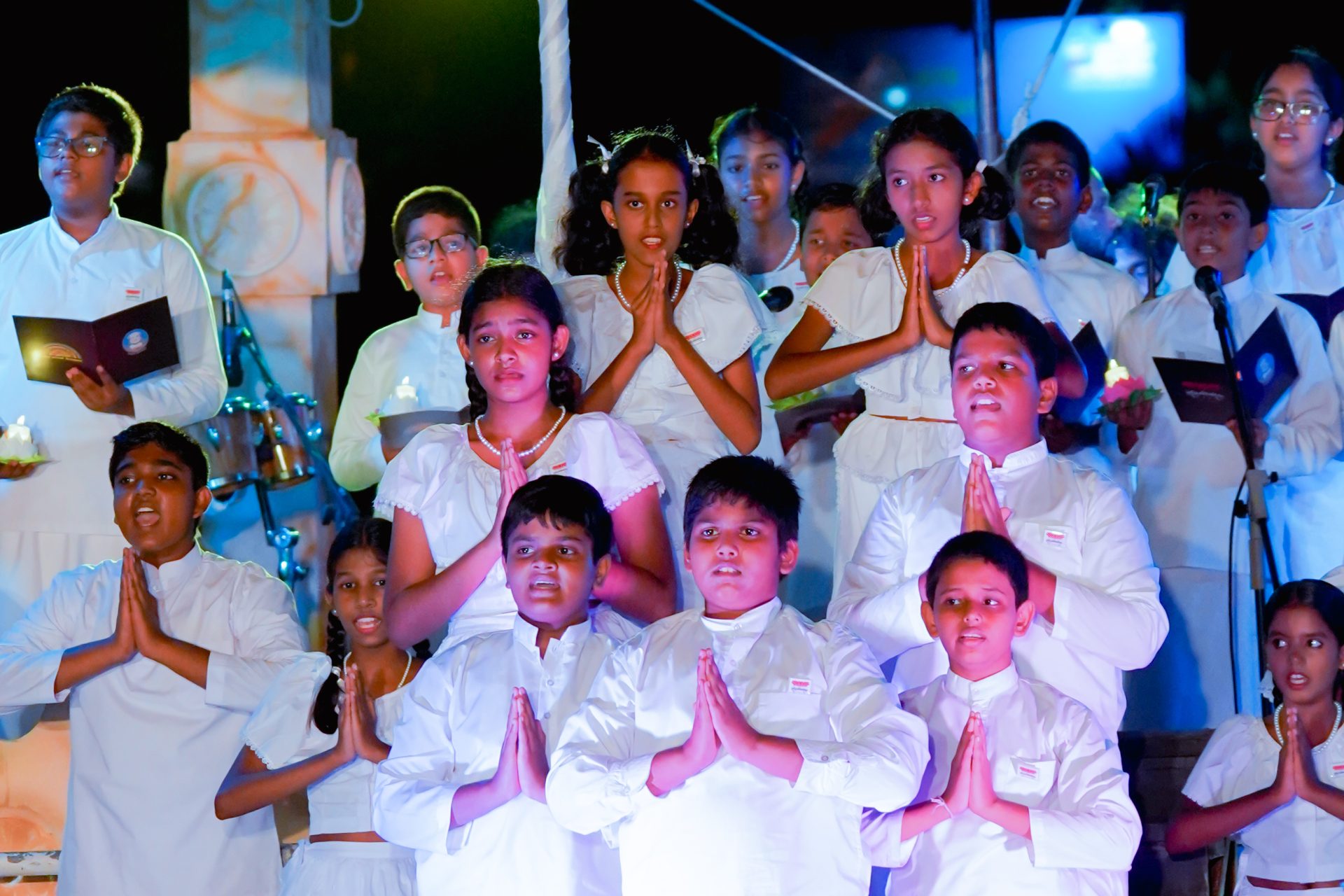 Children in white praying at a ceremonial event with candles and hymn books.
