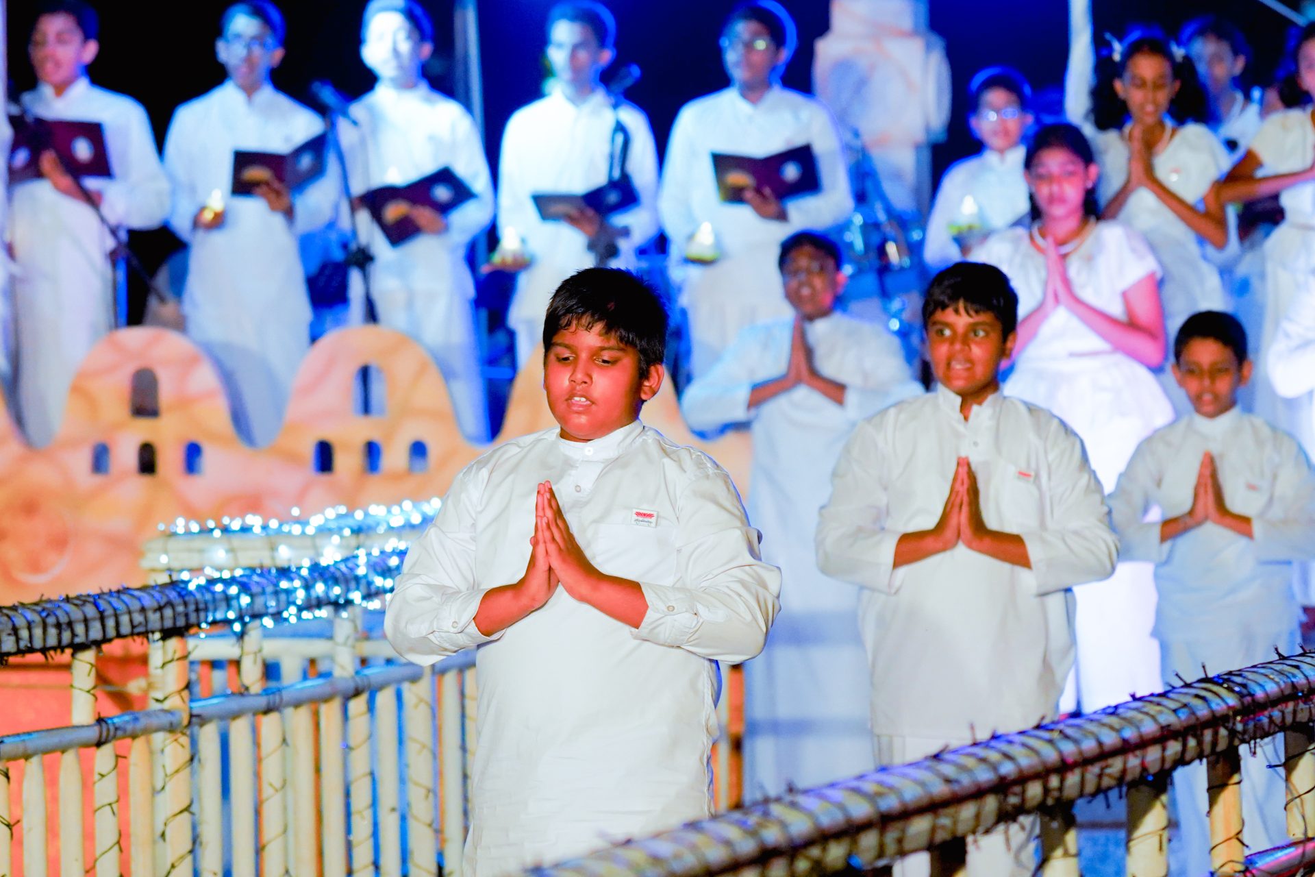 Children in white garments praying on lit bamboo bridge during ceremonial event.
