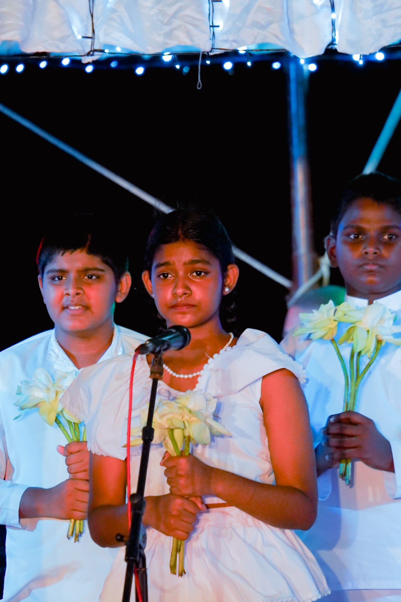 Children in white attire on stage with flowers during solemn ceremony.
