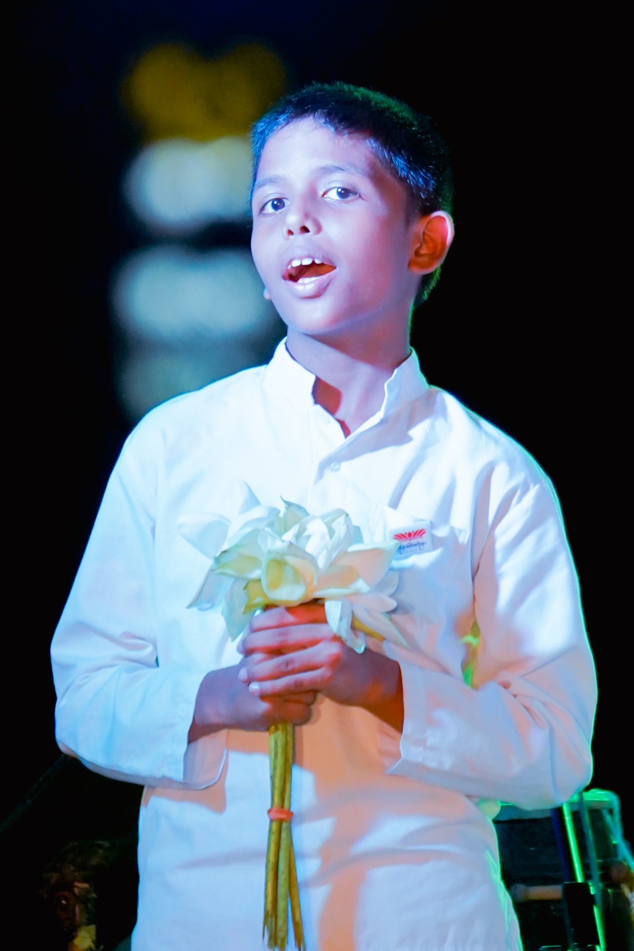 Young boy singing on stage with flowers, illuminated by stage lights.