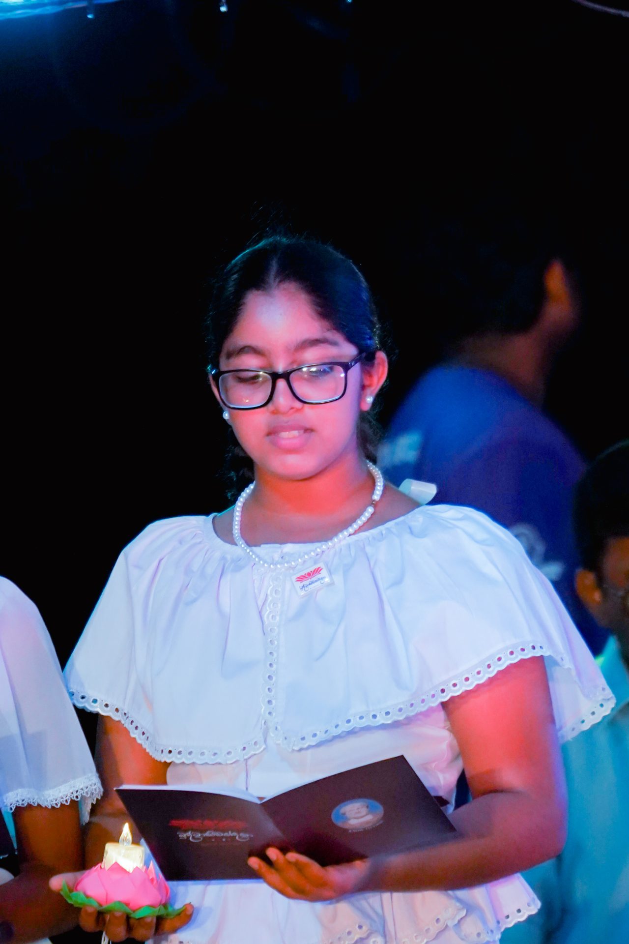 Young girl in white dress reading booklet, holding lit candle at formal ceremony.