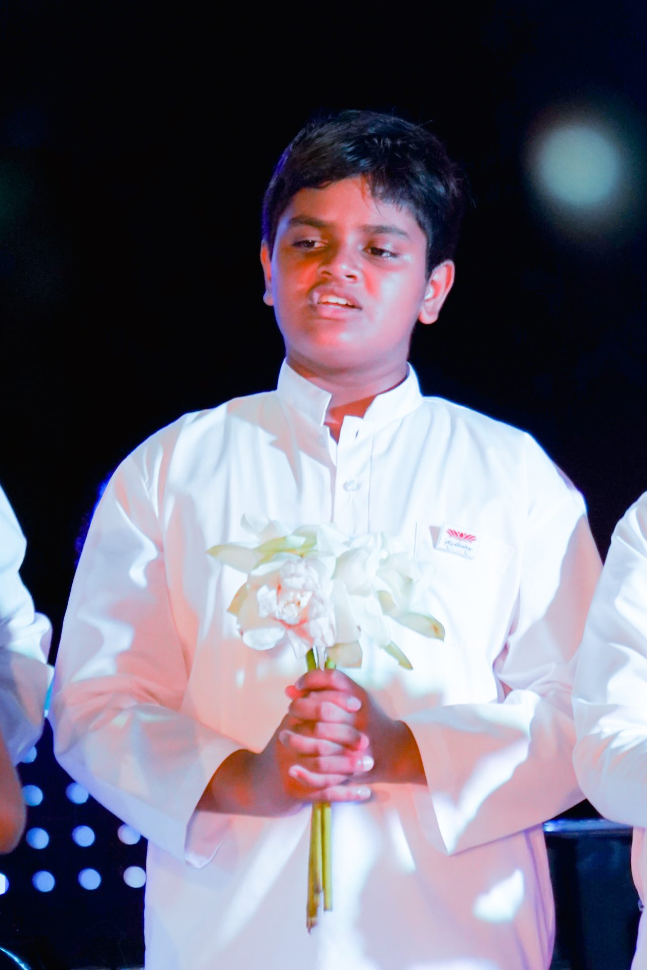 Young boy in white garment holding white flowers during a ceremony.