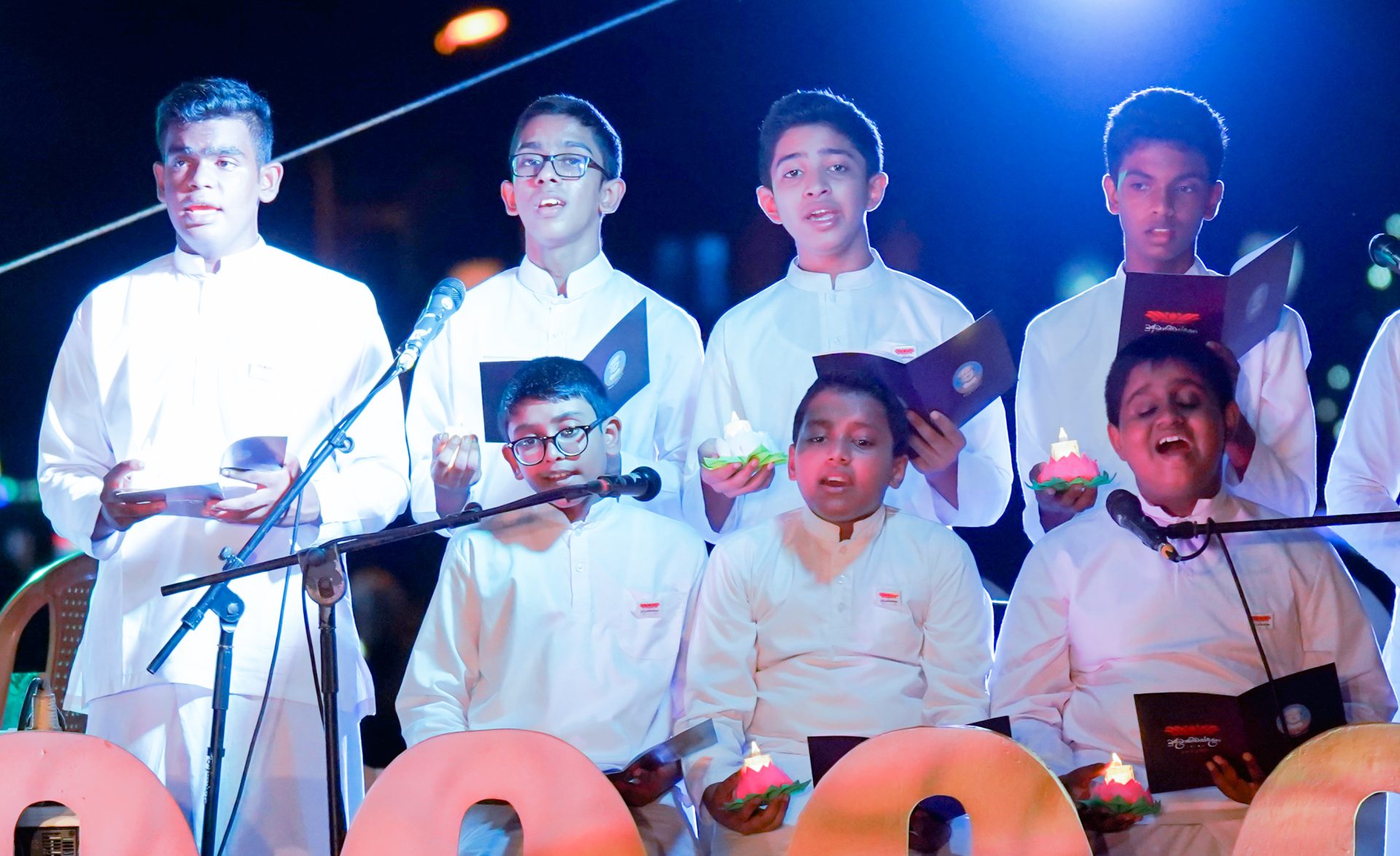 Boys choir in white attire performing on stage with dramatic lighting.