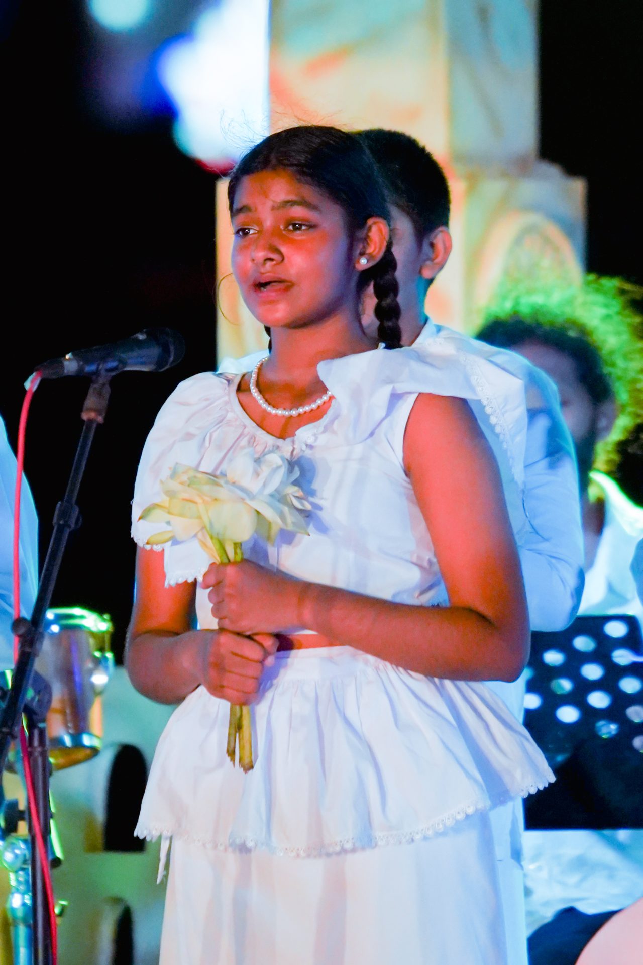 Young girl singing, holding flowers, in a formal performance setting.