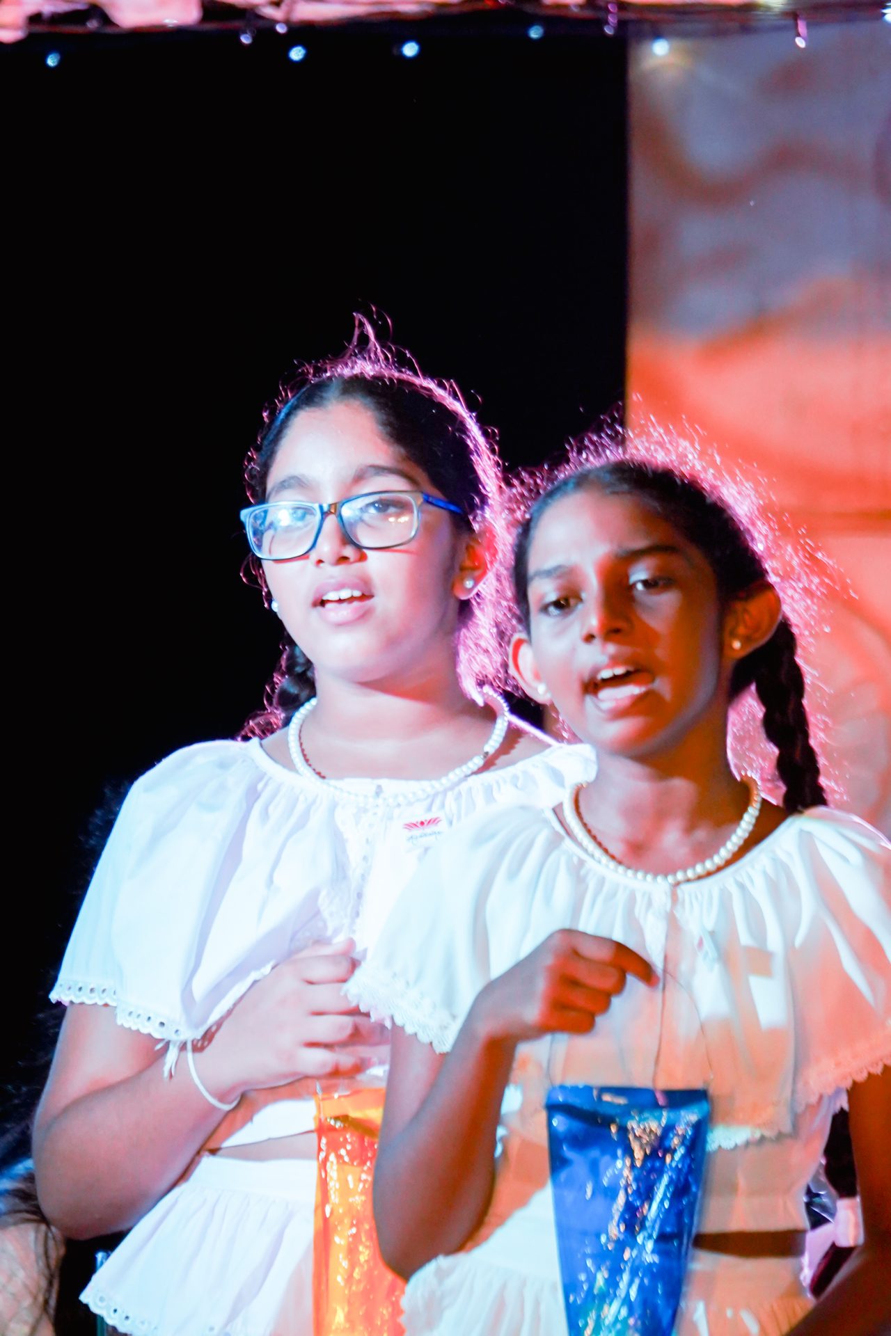 Two young girls singing in traditional white blouses during a festive school performance.