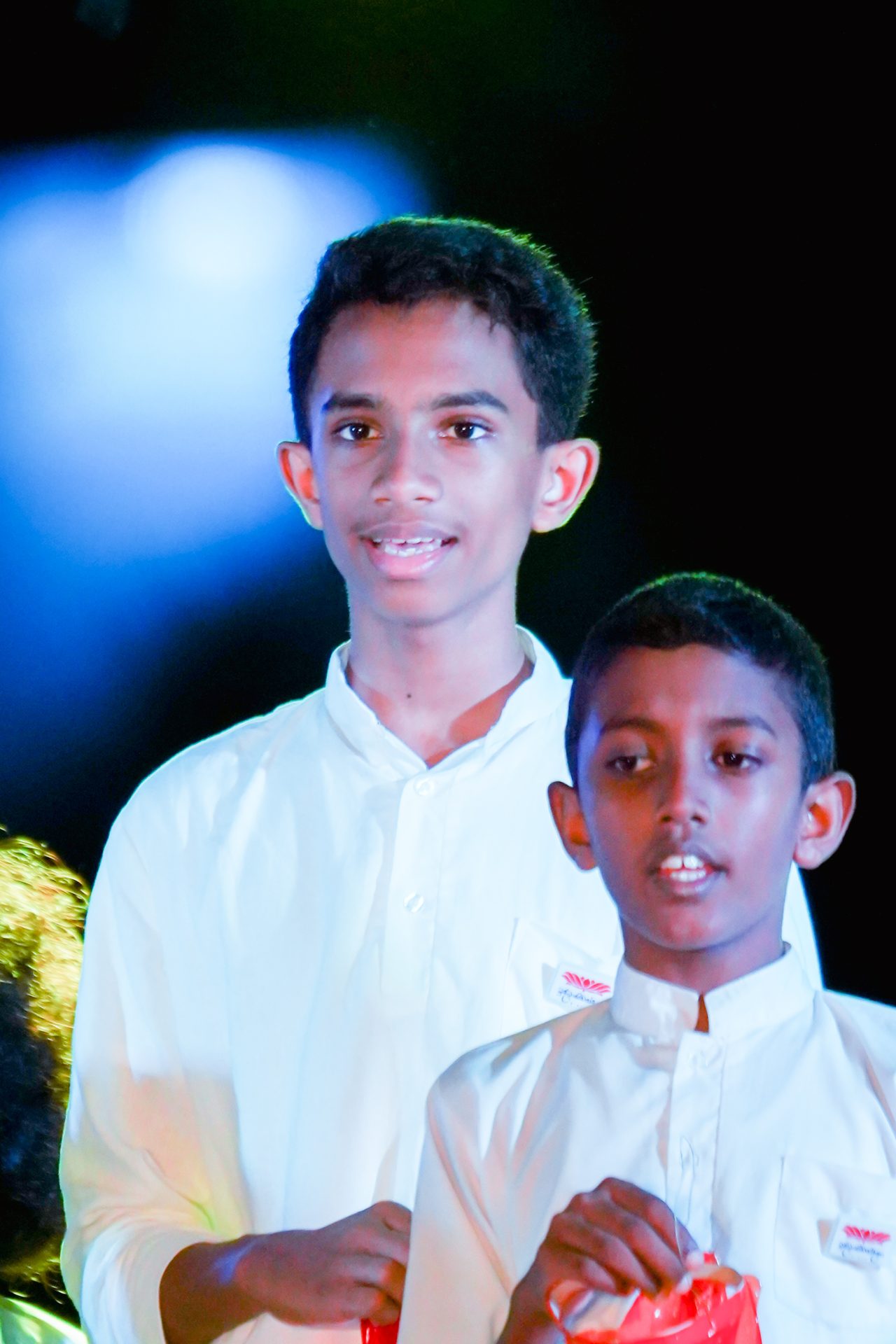 Two boys in white shirts holding red bags under colorful stage lighting.