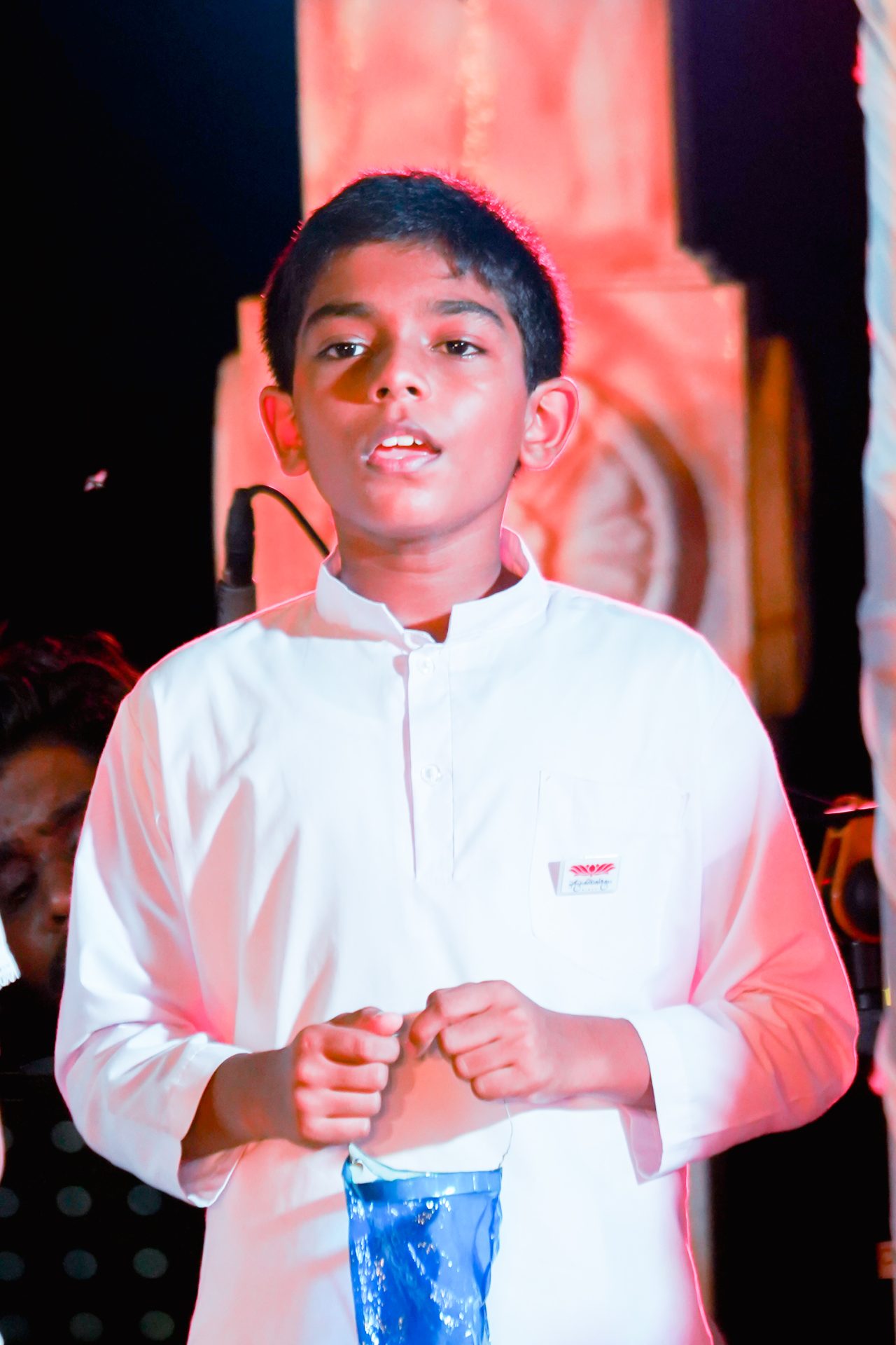 Young boy in white traditional attire holding a blue face mask on stage.