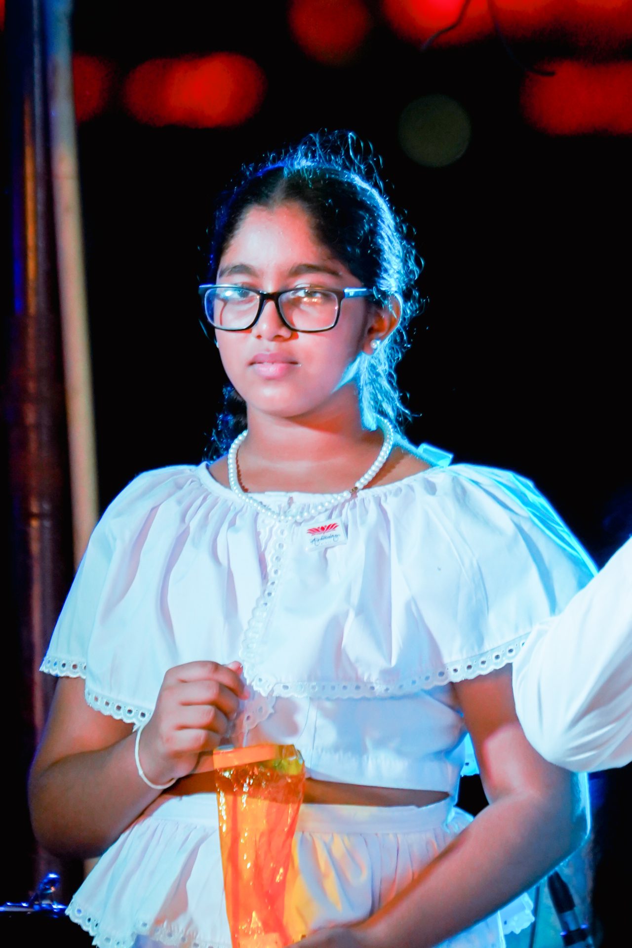 Young girl in traditional white outfit holding an orange item, festive background.