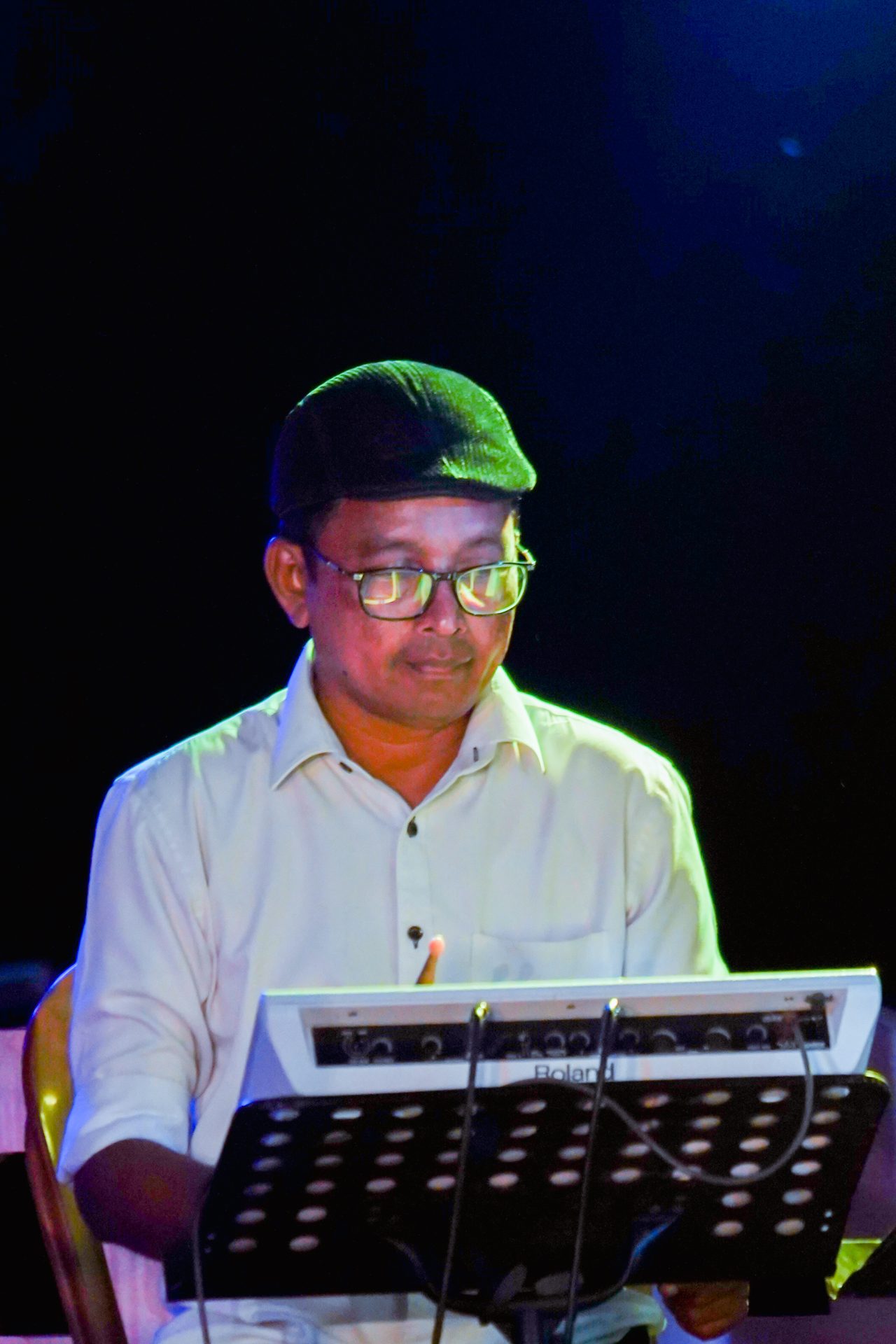 Musician in glasses playing Roland keyboard under spotlight on dim stage.