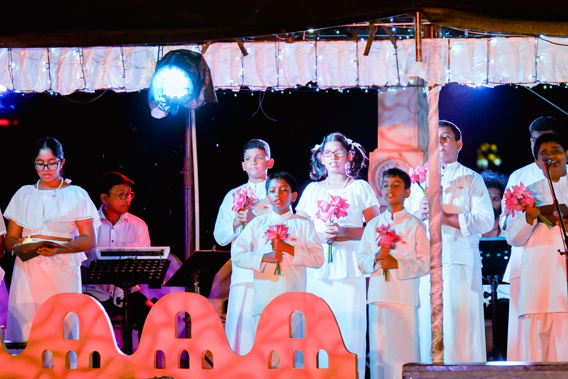 Children in white outfits performing on stage with flowers and music.
