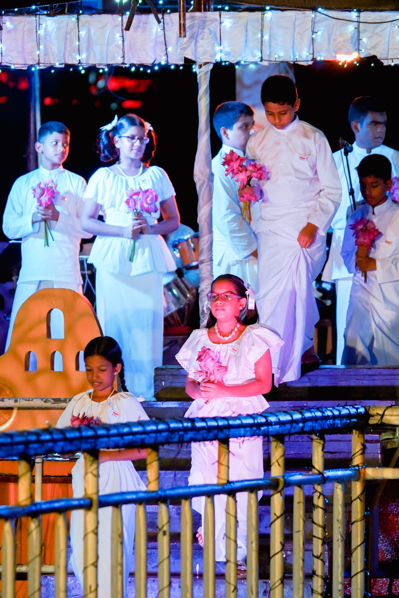 Children in white attire holding pink flowers at a ceremonial event.