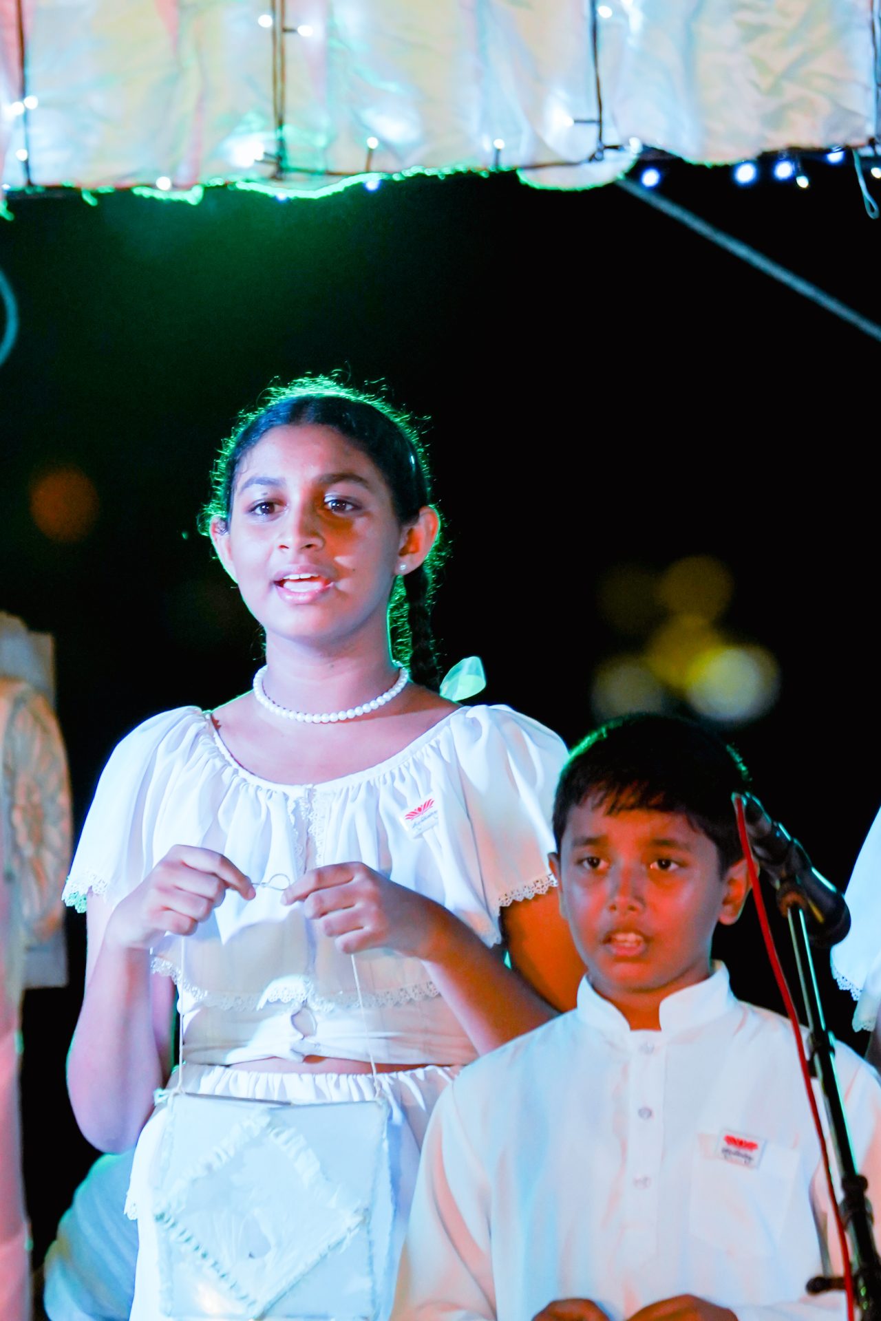 Children performing in white outfits with microphone and twinkling lights.