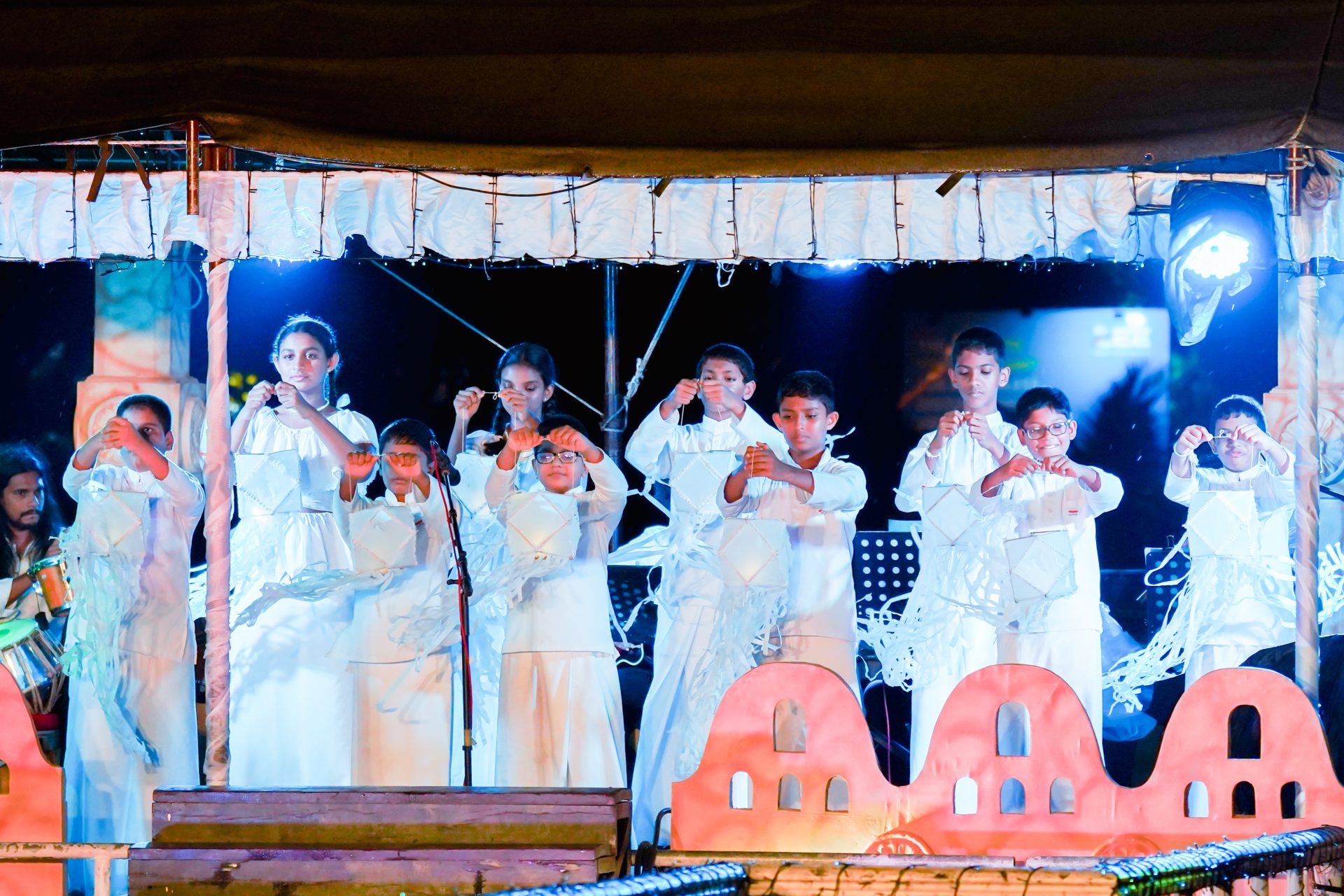 Children holding white lanterns on stage during a cultural ceremony.