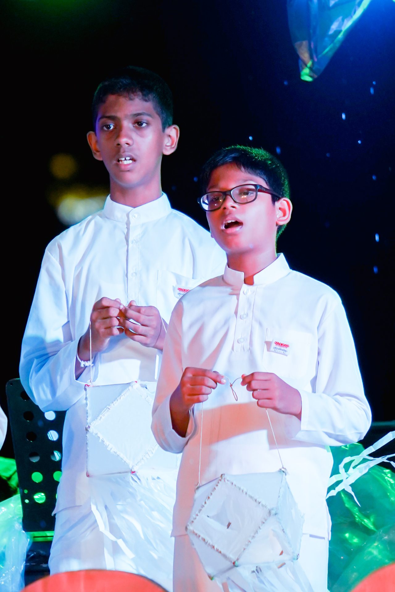 Two boys holding lanterns during an evening ceremony, dressed in traditional white attire.