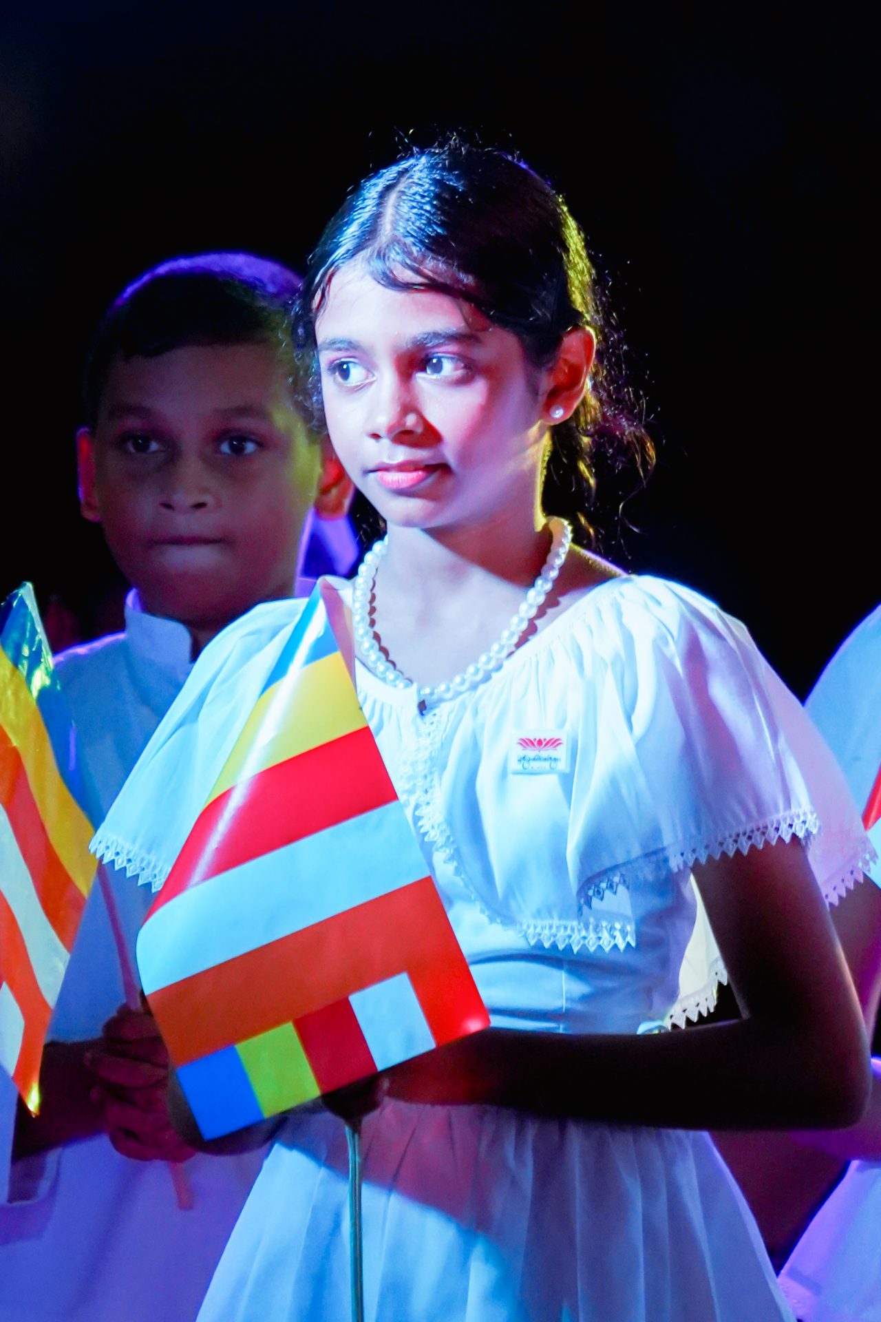 Children in traditional attire holding cultural flags during a formal ceremony.