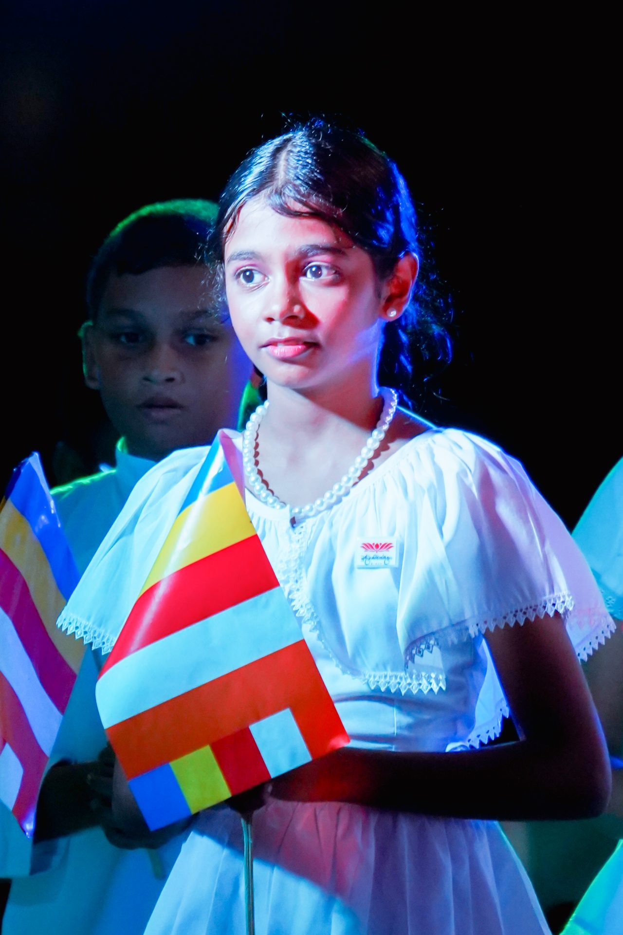 Young girl holding colorful flag in white dress with boy in background.
