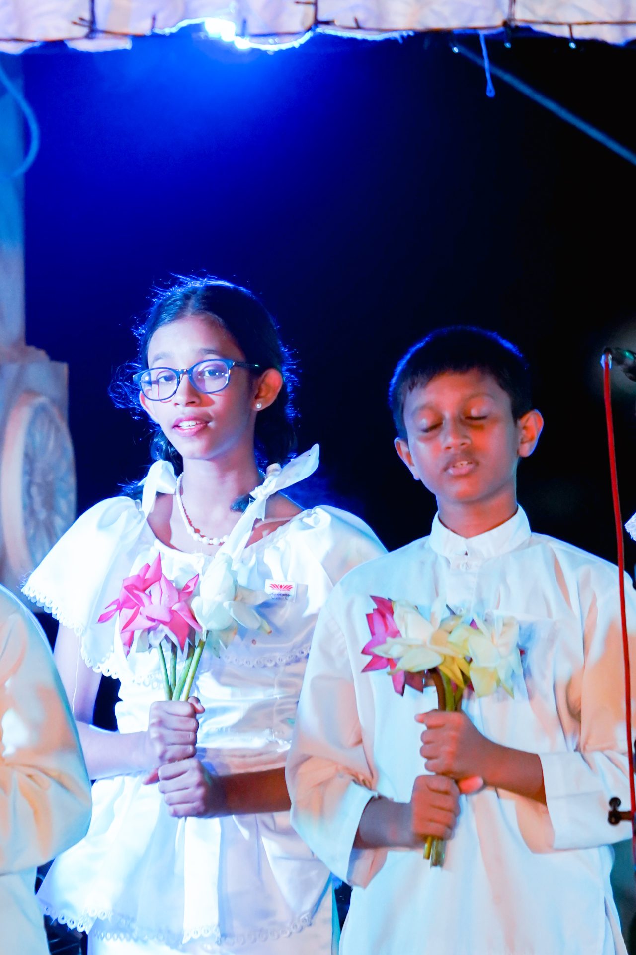 Children in white attire with flowers under blue stage light during a formal ceremony.