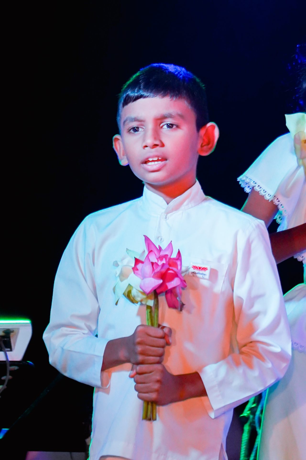 Boy holding pink lotuses on stage during a ceremonial event.