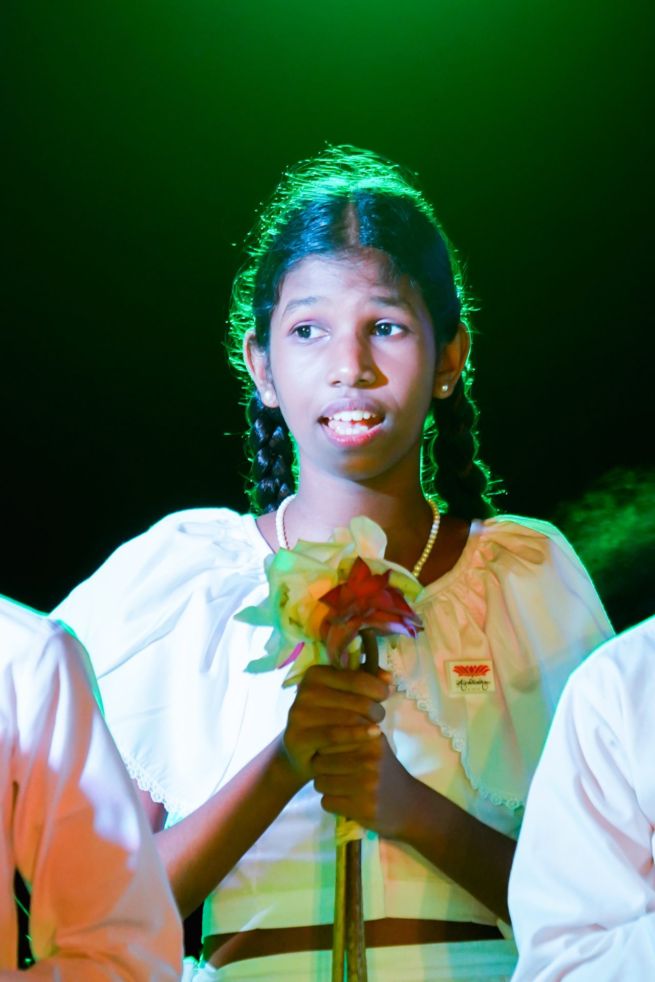 Young girl in traditional white outfit holding a bouquet on stage.