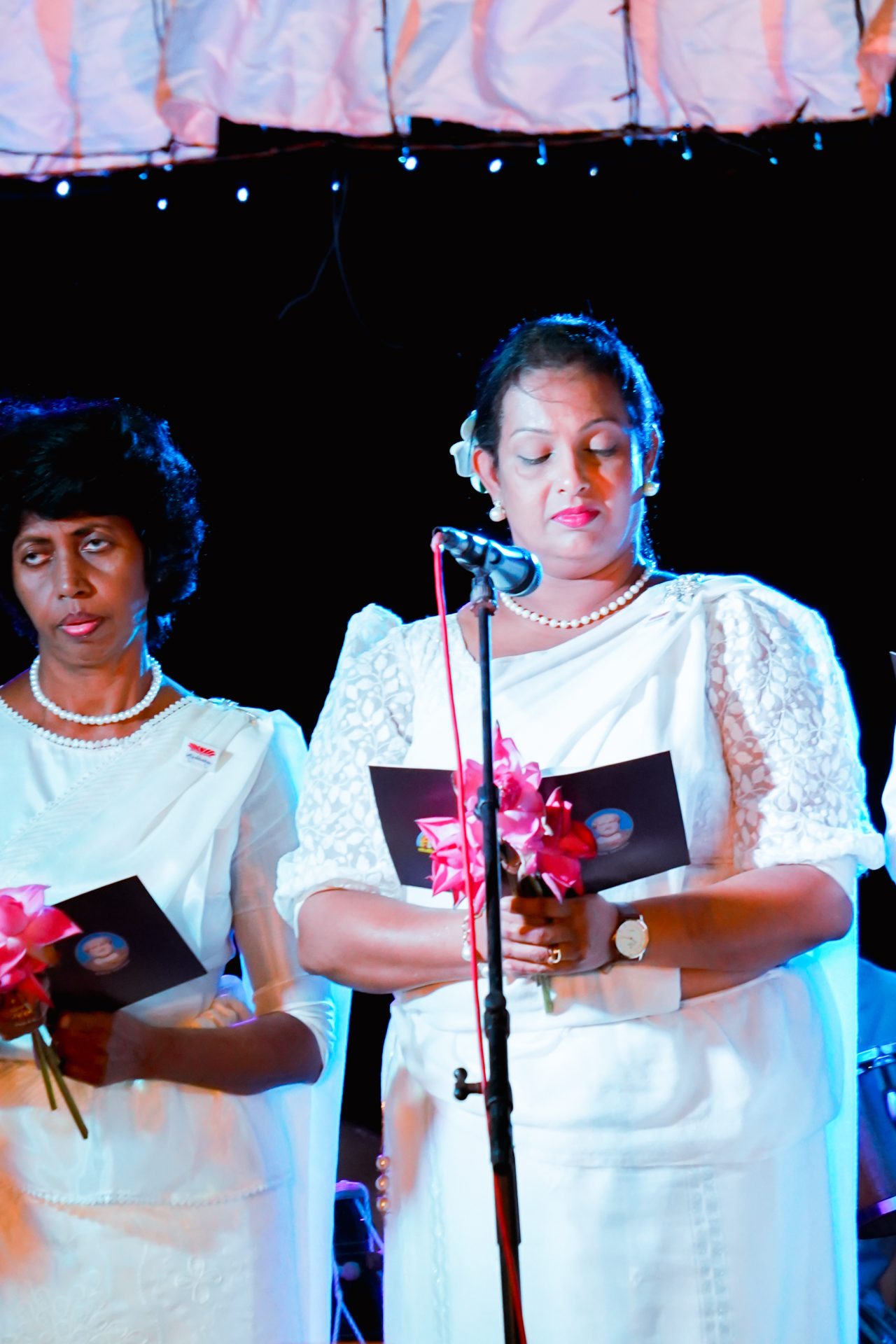 Two women in white lace saris at a formal event, holding booklets and pink flowers.