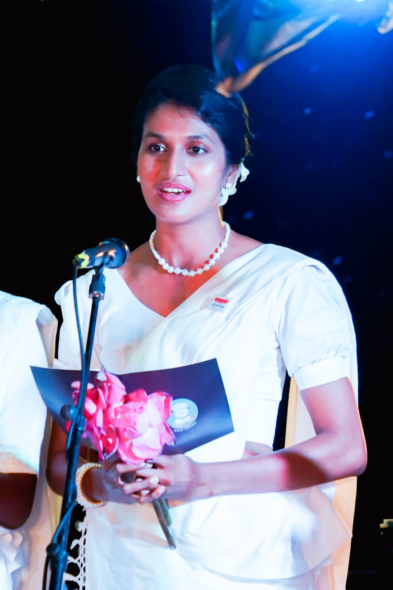 Young woman in white saree speaking at formal event.